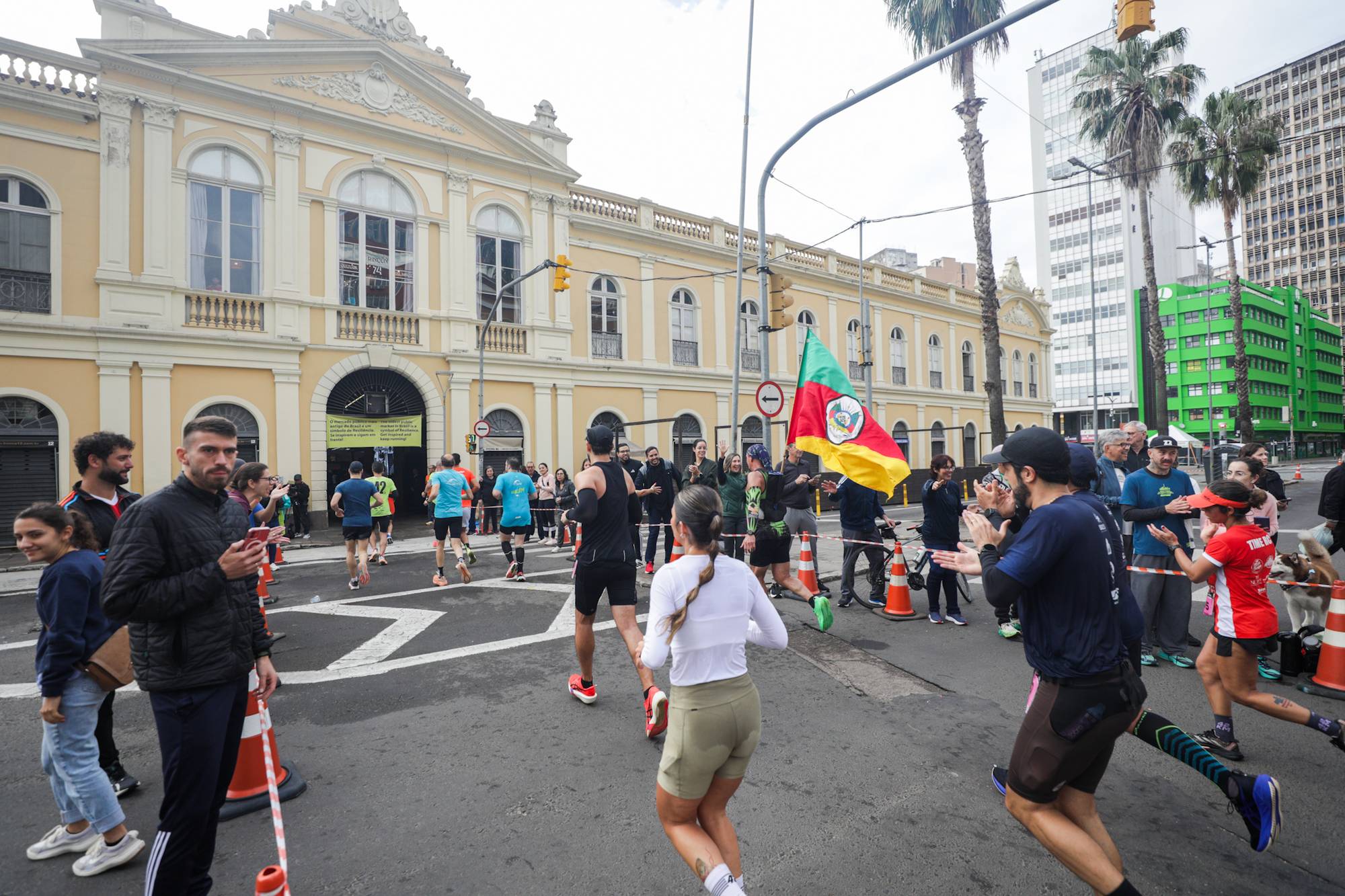 Corredores com número de peito passando em frente ao Mercado Público de Porto Alegre, com torcedores aplaudindo na calçada, um homem filmando com o celular e uma bandeira do Rio Grande do Sul erguida no meio da rua.
