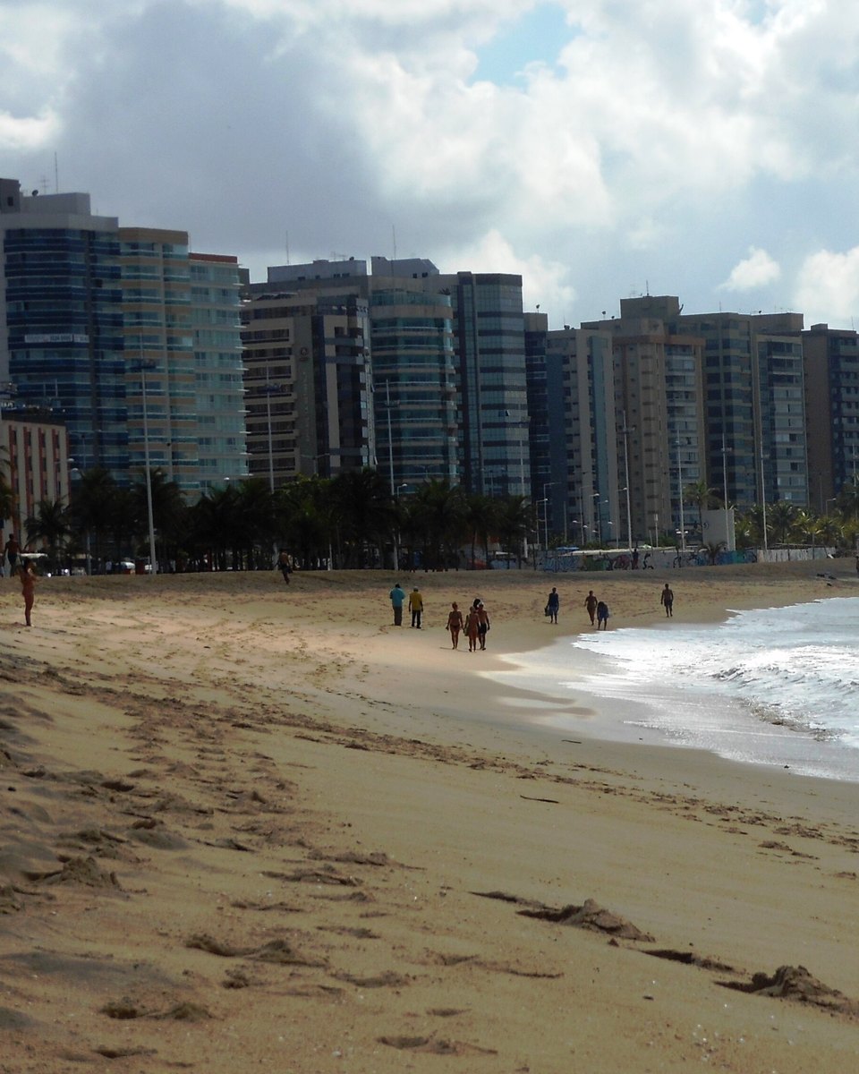 Pessoas caminhando na areia da Praia de Camburi, em Vitória, com a fileira de prédios da orla e as ondas ao lado.