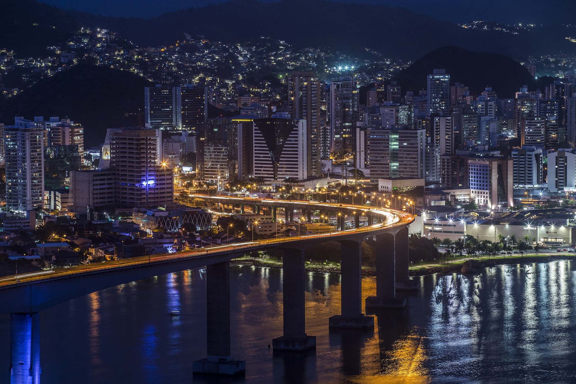 Terceira Ponte iluminada à noite sobre a baía, com os prédios e as luzes de Vitória ao fundo e o reflexo na água.