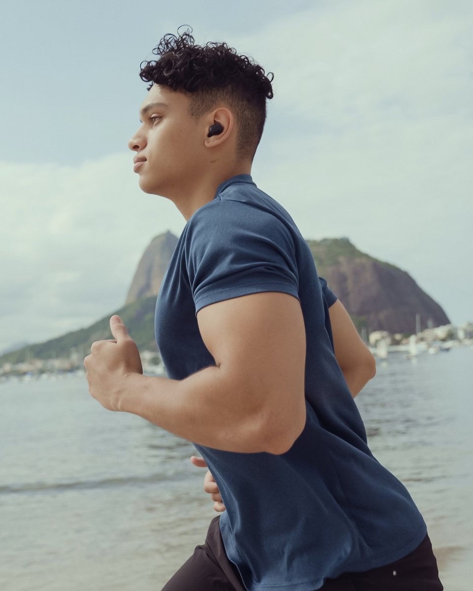 Jovem de camiseta azul e fone sem fio correndo na areia da Baía de Guanabara, no Rio de Janeiro, com o Pão de Açúcar e barcos ancorados ao fundo.