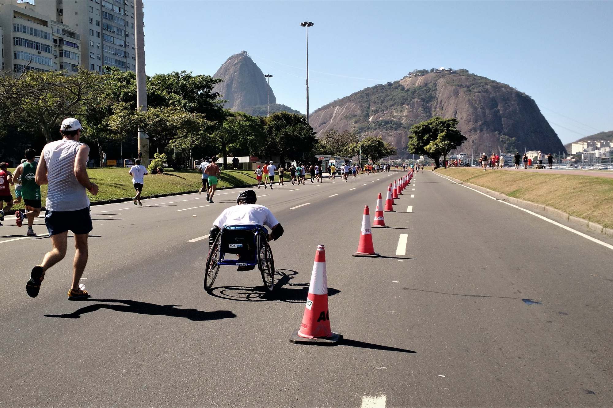 Corredores e um atleta em cadeira de rodas numa pista do Aterro do Flamengo, no Rio de Janeiro, com cones laranja, árvores e o Pão de Açúcar ao fundo num dia de céu azul.