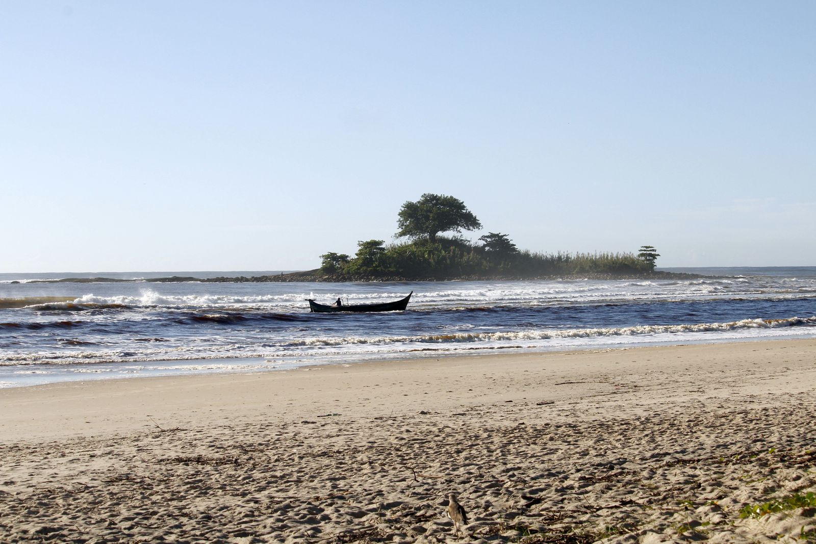 Praia de Guaratuba, no litoral do Paraná, com um pescador numa canoa entre as ondas, uma ilhota coberta de árvores ao fundo e a areia em primeiro plano, numa manhã de céu limpo em março.