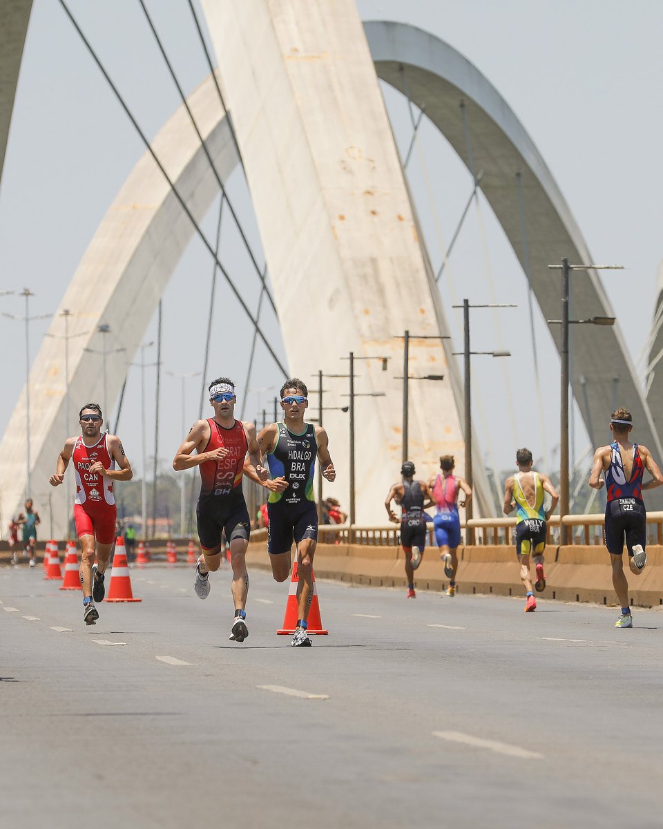 Atletas correndo em fila na pista da ponte de três arcos sobre o Lago Paranoá, em Brasília, com cones laranja na faixa e uma moto de apoio à frente, durante a etapa de Brasília da Copa do Mundo de Triathlon de 2023.