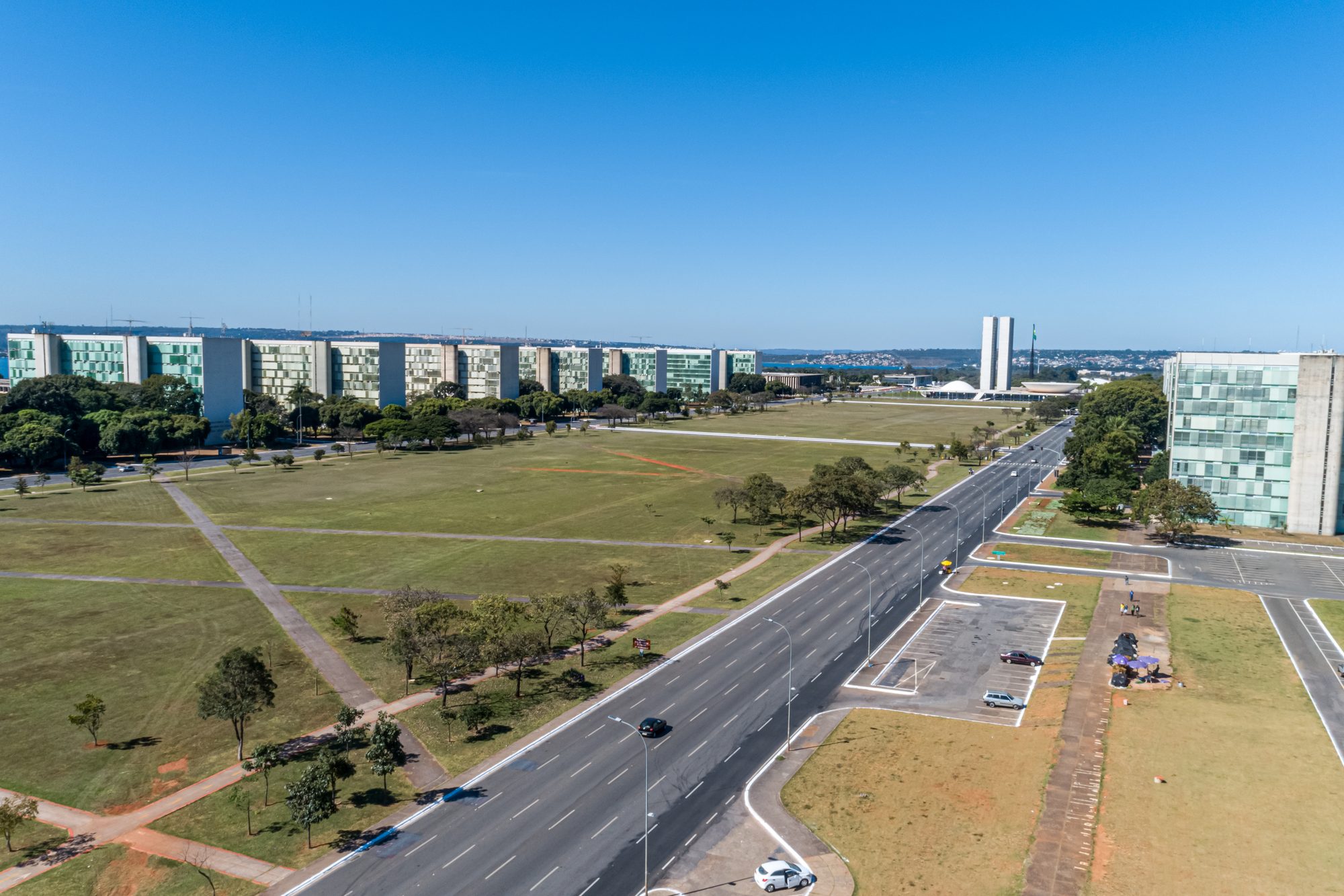 Vista do alto da Esplanada dos Ministérios, em Brasília, com o gramado central, a avenida de várias faixas em linha reta, a fila de prédios dos ministérios dos dois lados e o céu azul sem nuvens.