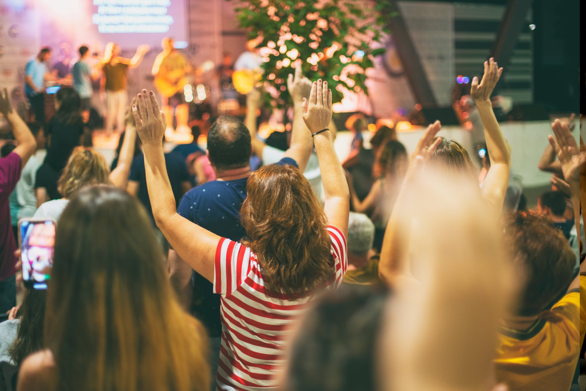 Público de mãos levantadas durante um louvor com banda no palco, com uma pessoa filmando com o celular.