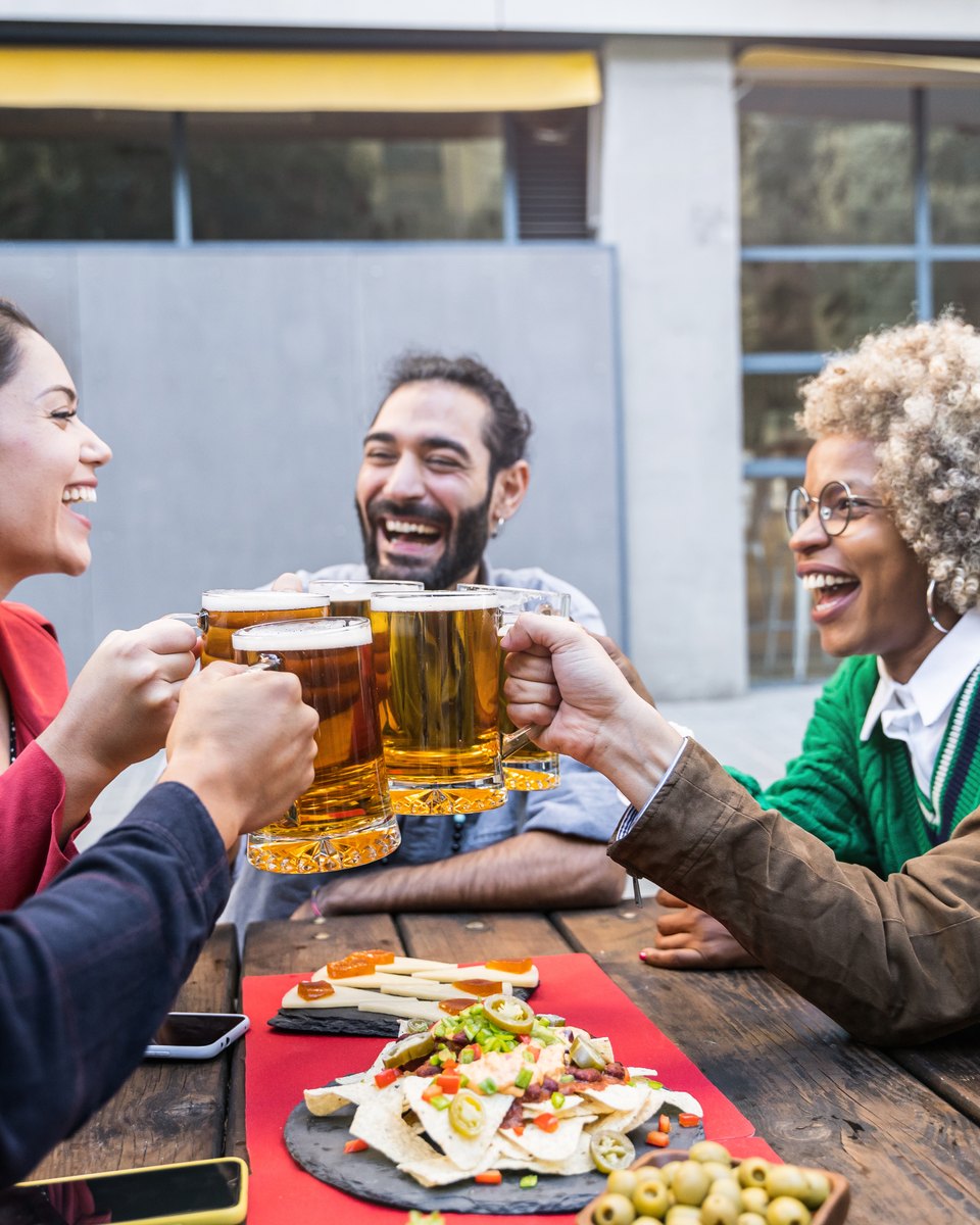 Cinco amigos adultos rindo e brindando com canecas de cerveja numa mesa de madeira ao ar livre, com nachos e azeitonas no centro.