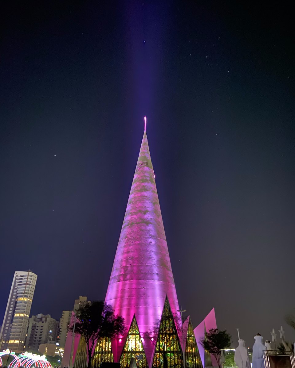 Catedral de Maringá iluminada de rosa numa noite de dezembro, com arcos de luzes de Natal à esquerda, figuras brancas de presépio à direita e visitantes na praça.