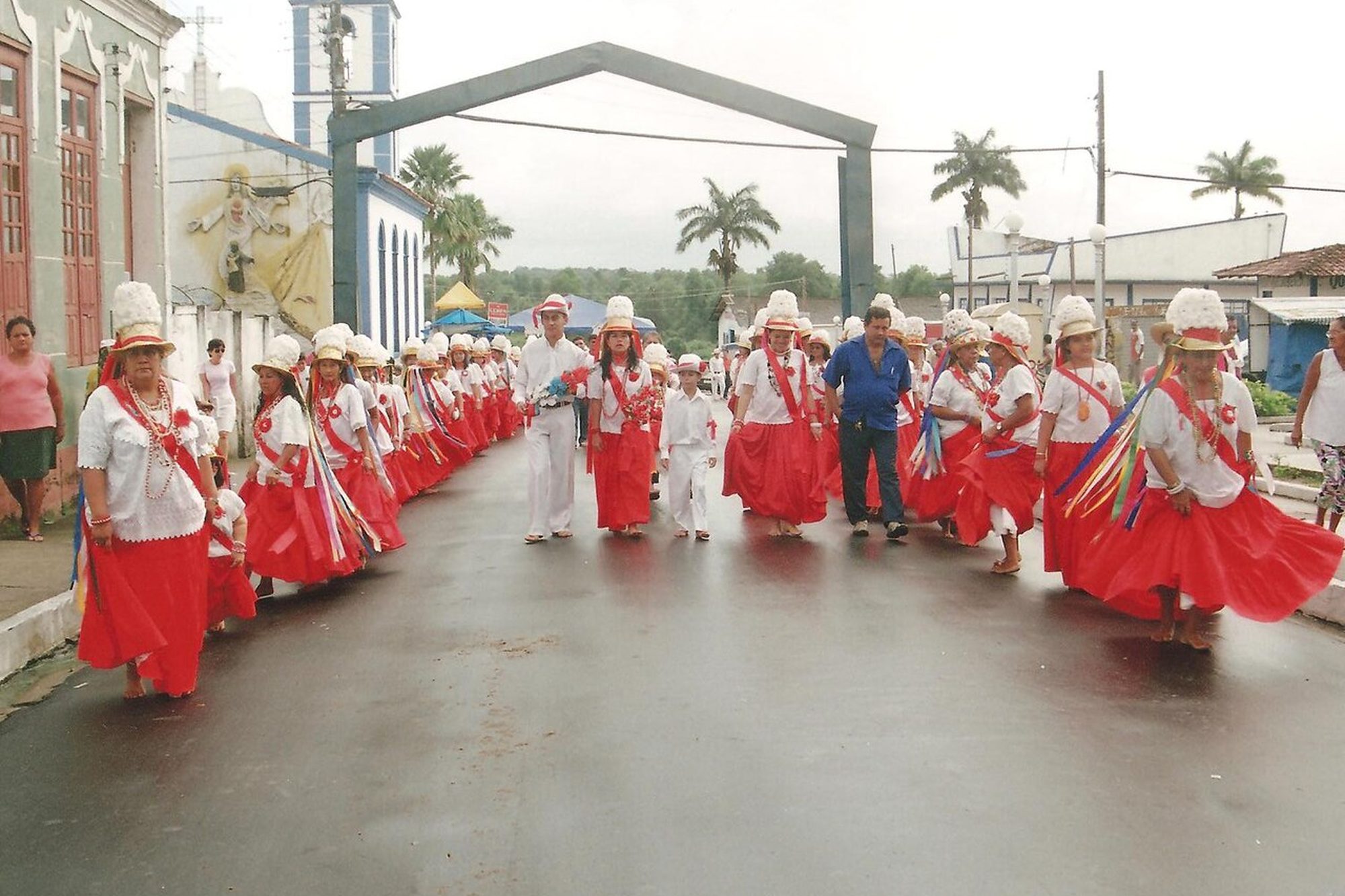 Marujas de saia vermelha rodada, blusa branca e chapéu de plumas e fitas caminhando por uma rua de Bragança, no Pará, com a igreja e palmeiras ao fundo.