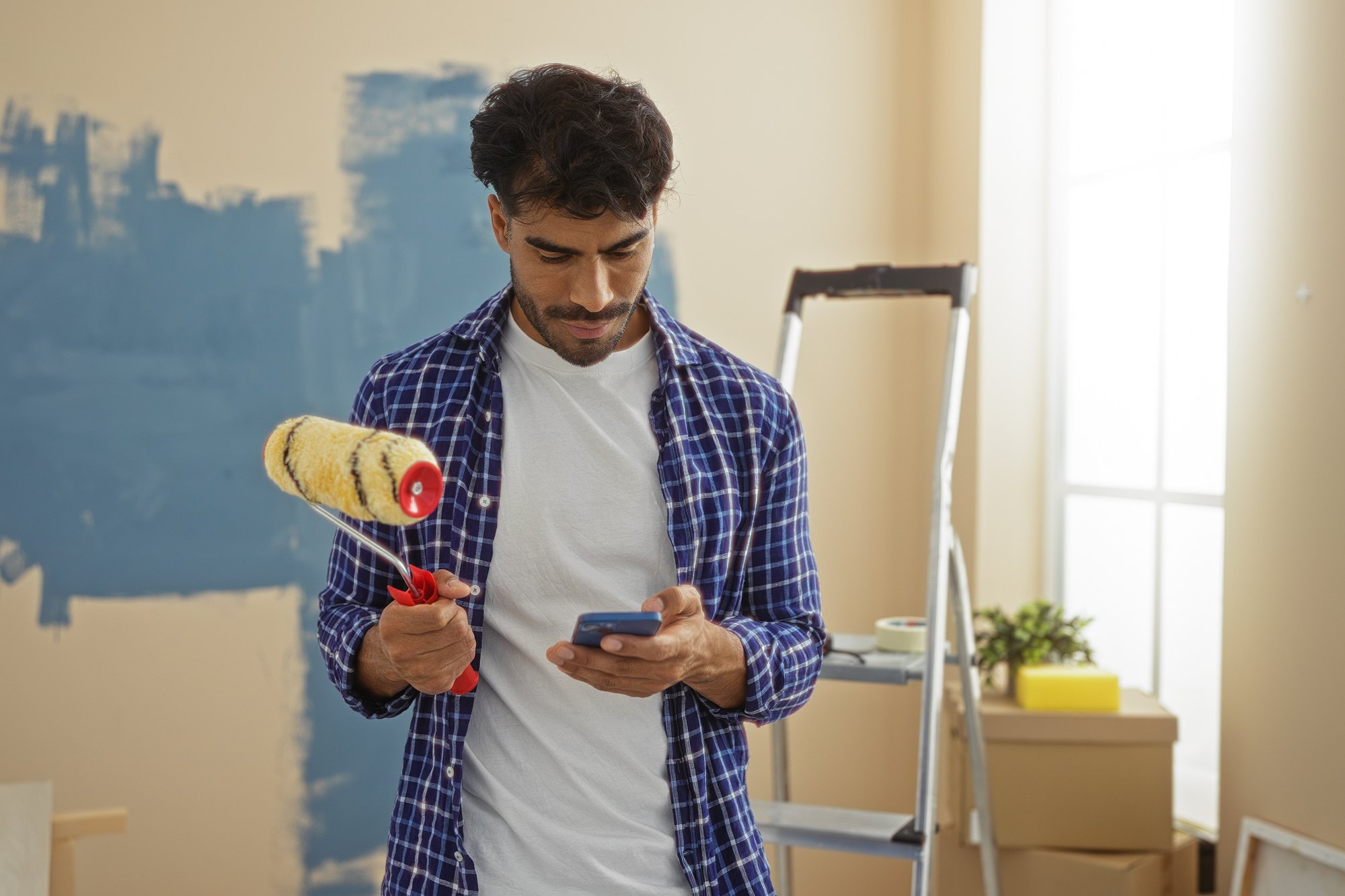 Homem de camisa xadrez segurando um rolo de pintura numa mão e o celular na outra, diante de uma parede pintada de azul pela metade, com escada e caixas de mudança ao fundo.