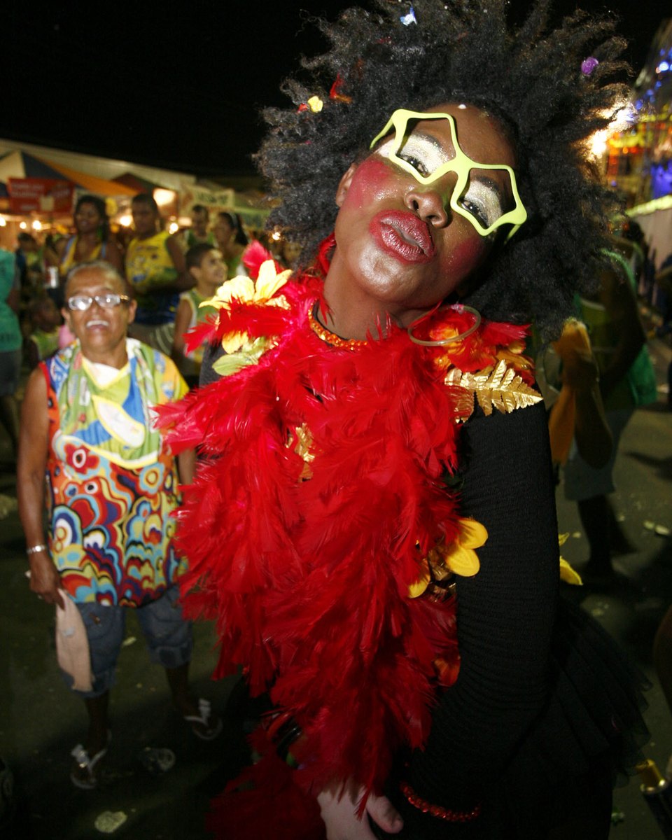 Foliã de cabelo black power com óculos amarelo em forma de estrela, maquiagem prateada, batom vermelho e boá de plumas vermelhas posando à noite no circuito da Micareta de Feira de Santana, com outros foliões e barracas iluminadas ao fundo.