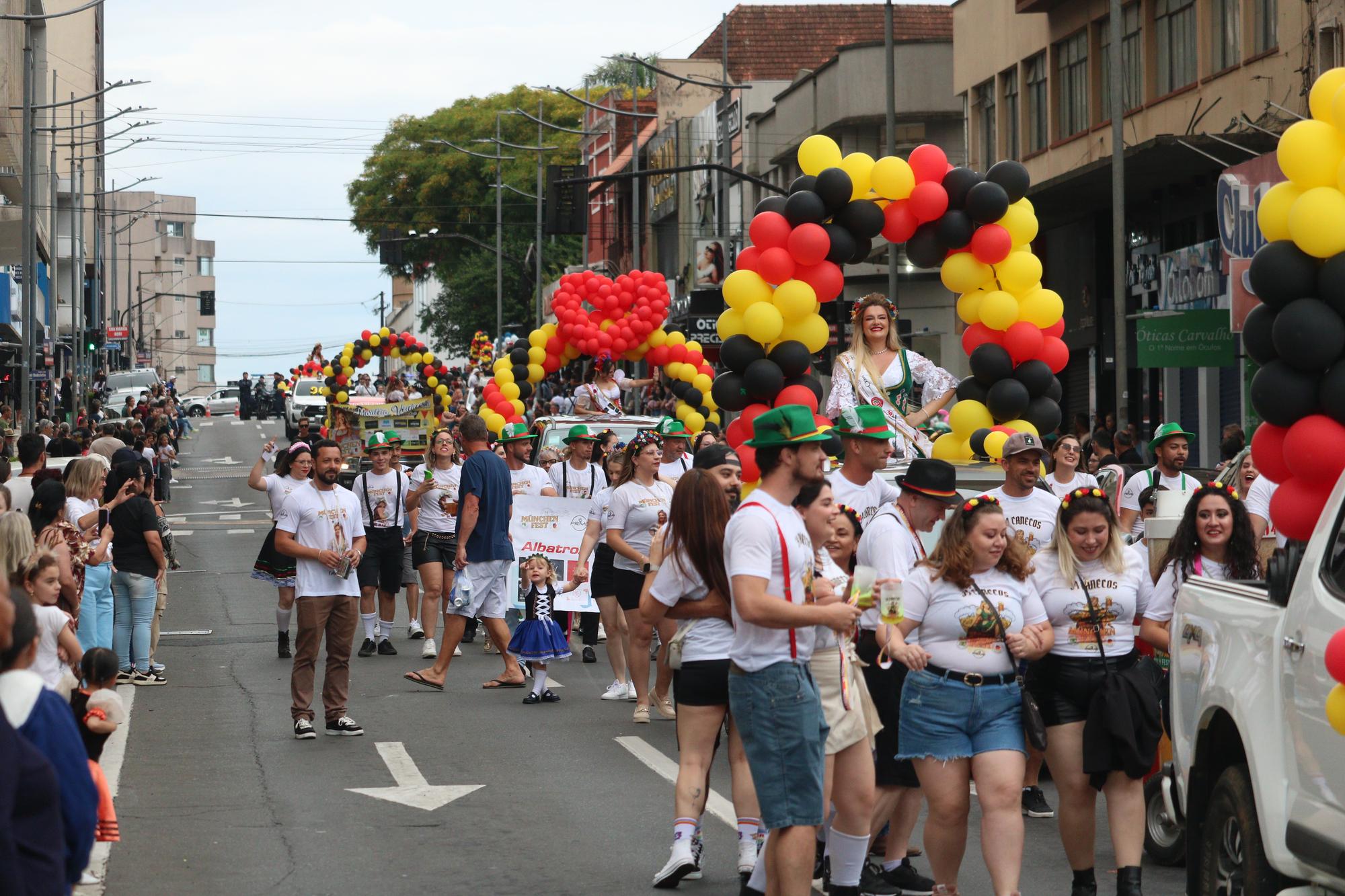 Desfile de blocos da Münchenfest na Avenida Vicente Machado, em Ponta Grossa: participantes com chapéu tirolês verde, suspensório e camisetas do bloco caminham entre arcos de balões preto, vermelho e amarelo, com público na calçada e prédios do Centro ao fundo.