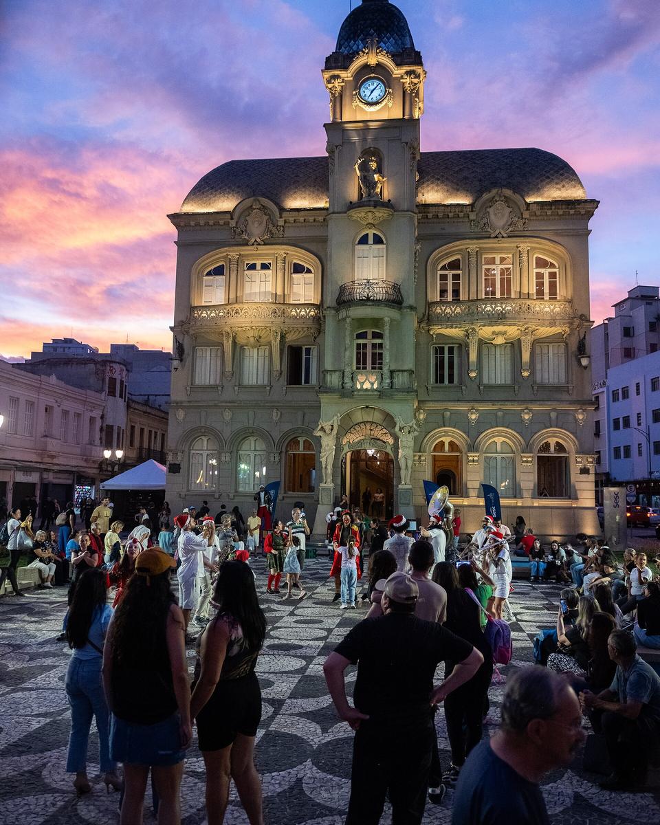 Público na Praça Generoso Marques, em Curitiba, assistindo a uma banda com gorros de Papai Noel diante do Paço da Liberdade iluminado, ao entardecer de dezembro, com o céu rosa e lilás atrás da torre do relógio.