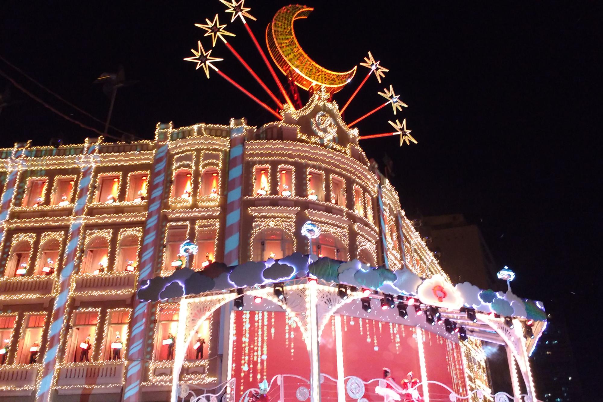Fachada do Palácio Avenida, na Rua XV de Novembro em Curitiba, coberta de luzes de Natal à noite, com o coral nas janelas iluminadas, uma lua dourada com estrelas no alto e o palco decorado com nuvens na frente.