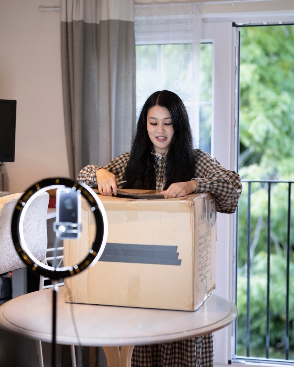 Mulher gravando um vídeo de unboxing em casa, abrindo uma caixa de papelão grande sobre uma mesa redonda, com o celular preso num tripé dentro de um ring light aceso.