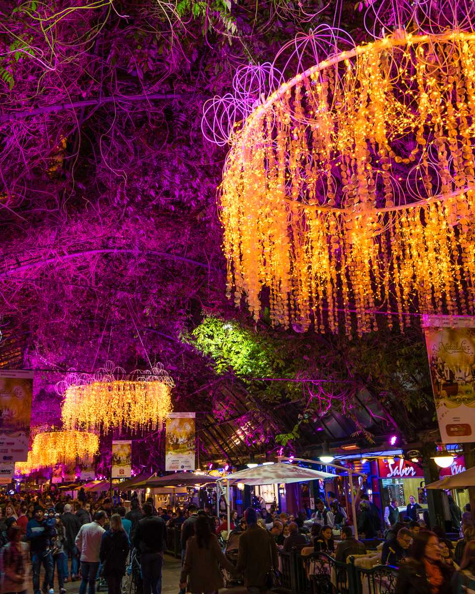 Rua Coberta de Gramado à noite durante o Natal Luz, com grandes lustres de luzes douradas pendurados sob as trepadeiras iluminadas de rosa e visitantes nas mesas dos cafés e restaurantes.