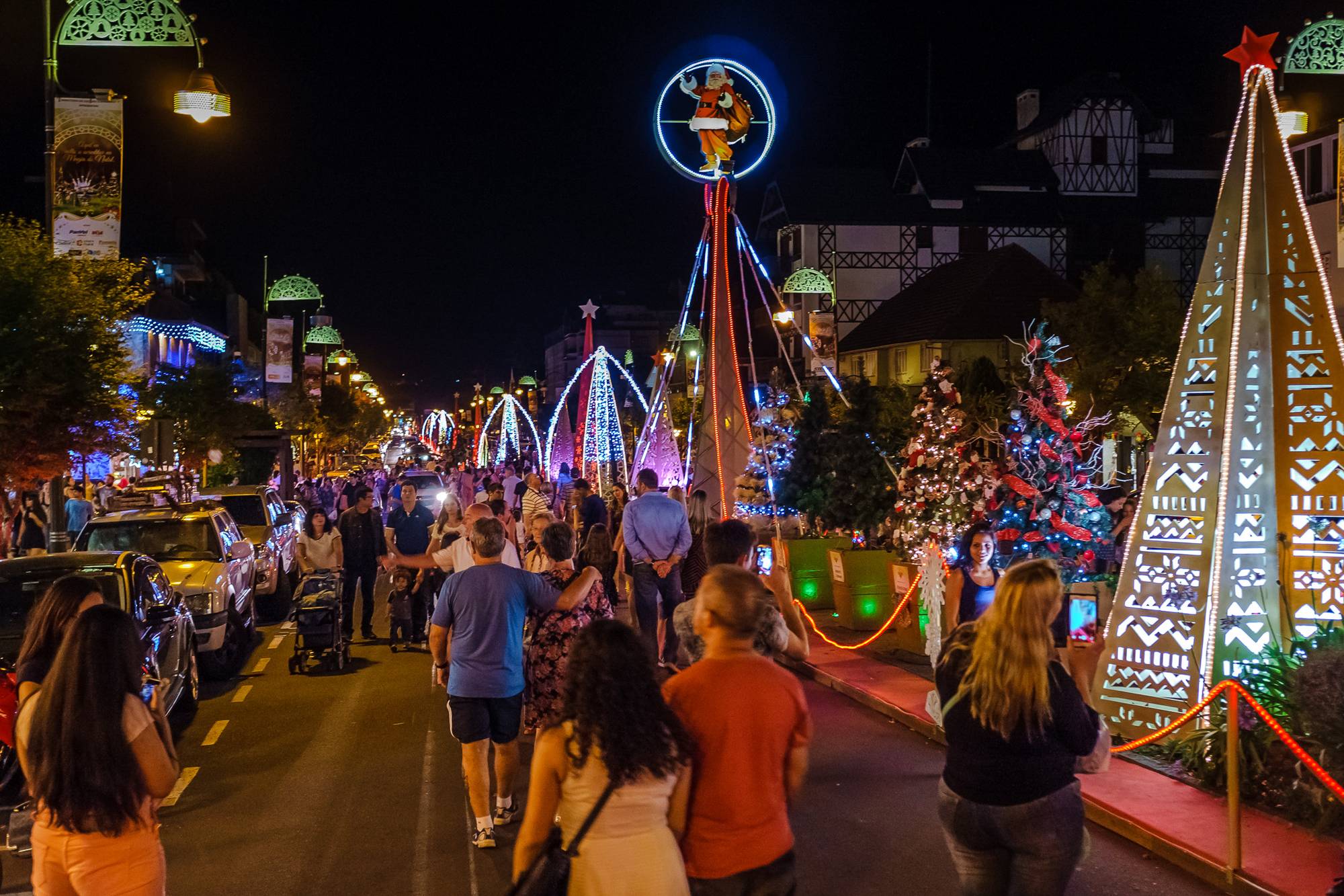 Público caminhando à noite pela Avenida Borges de Medeiros, em Gramado, durante o Natal Luz, entre árvores de Natal e pirâmides iluminadas, com um Papai Noel de luz no alto de uma torre e visitantes filmando com o celular.