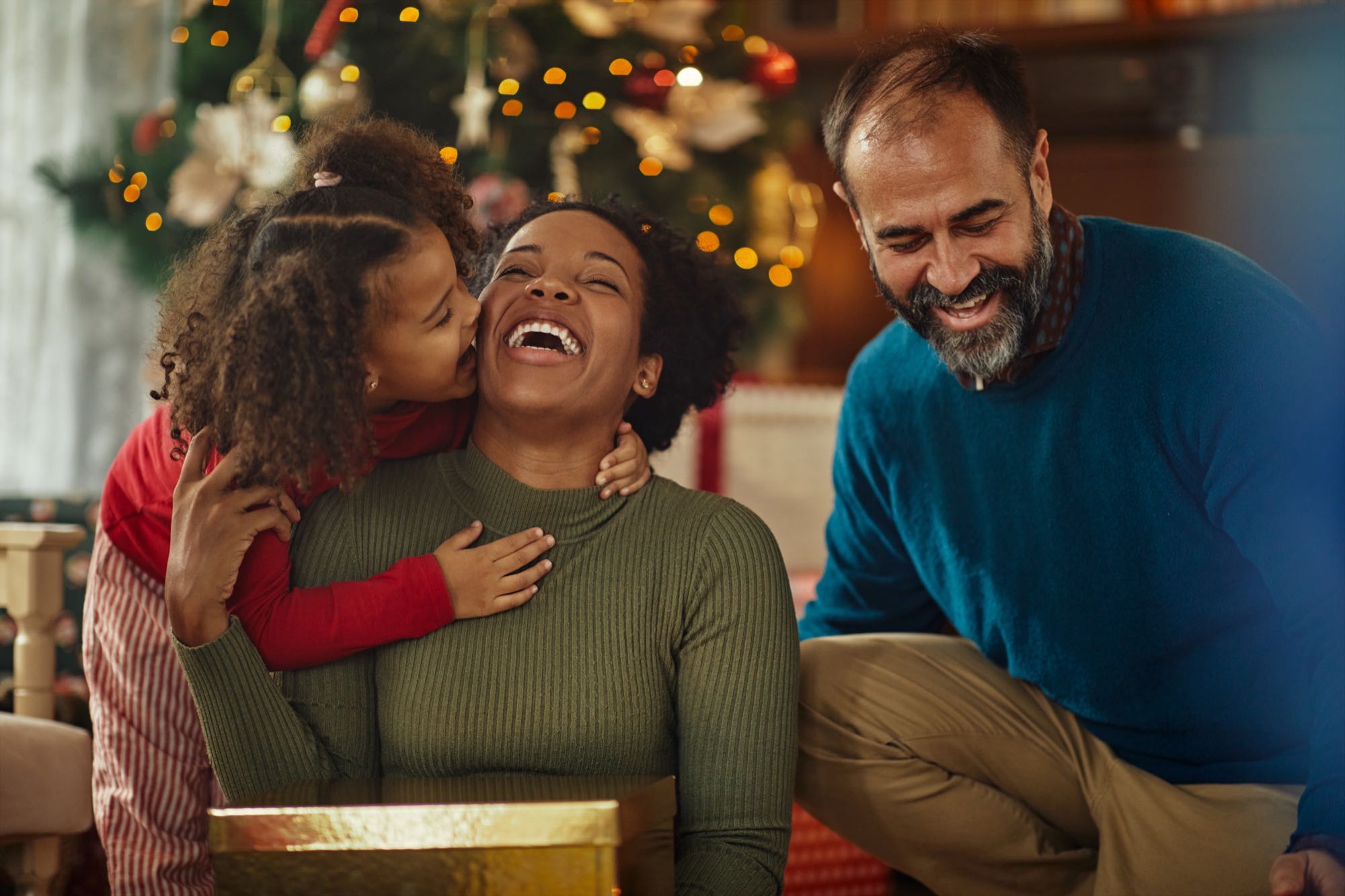 Família reunida no chão da sala diante da árvore de Natal, com uma menina abraçando e beijando a mãe que sorri e uma caixa de presente dourada aberta à frente.