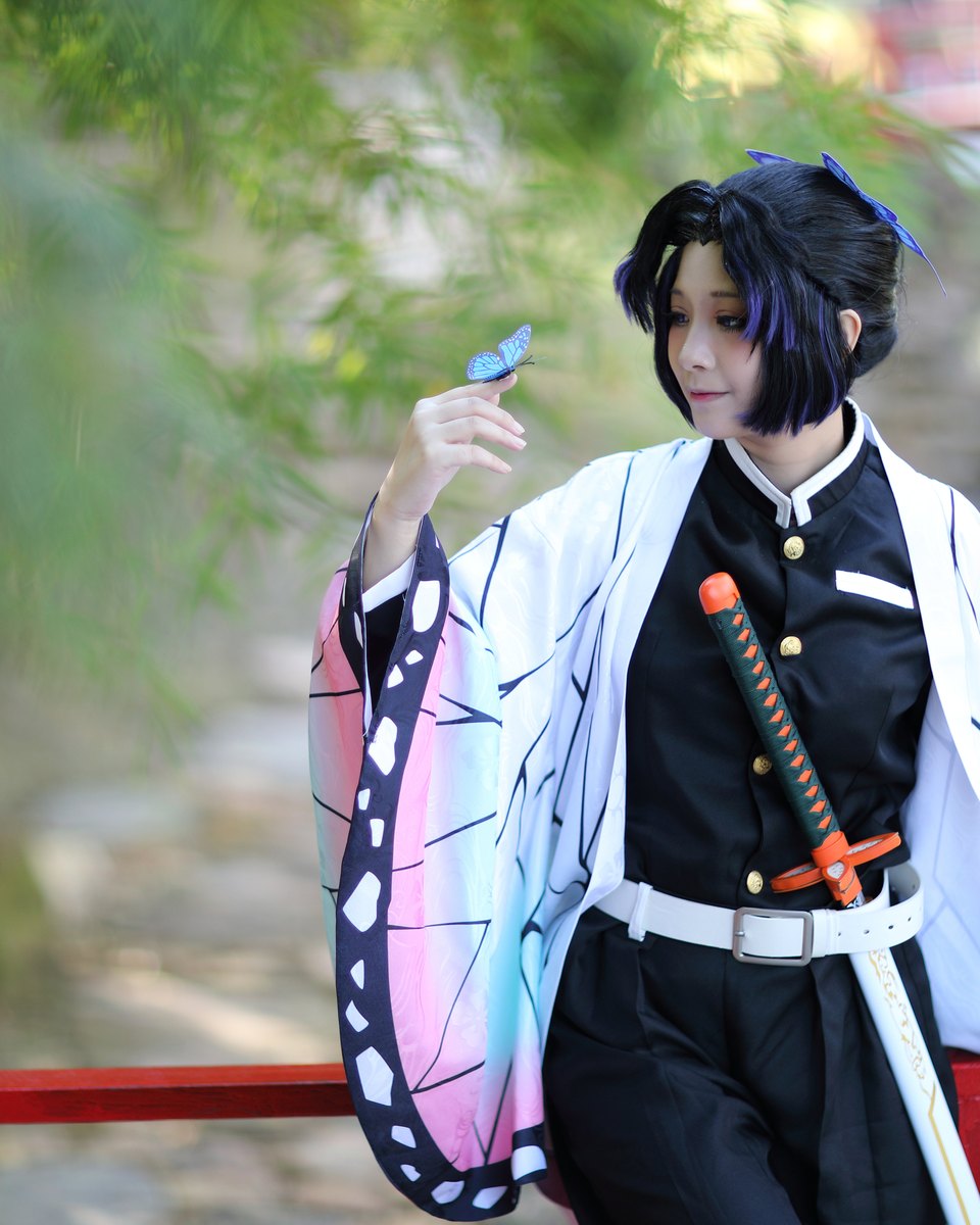 Cosplayer de peruca preta com mechas roxas, casaco branco estampado de asas de borboleta e espada na cintura, segurando uma borboleta azul de enfeite num jardim.