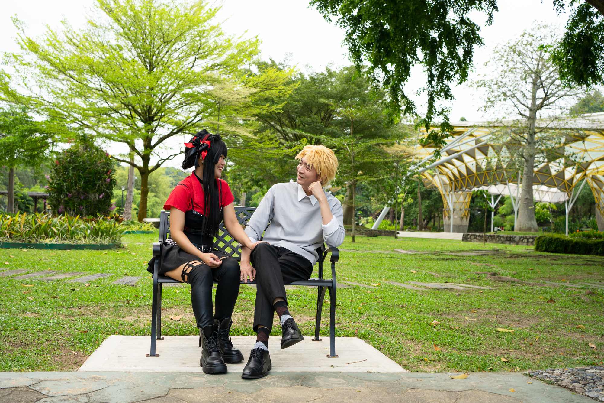 Dois cosplayers conversando e rindo sentados num banco de ferro num parque, ela de peruca preta com laço vermelho e bota de cano alto, ele de peruca loira e suéter cinza.
