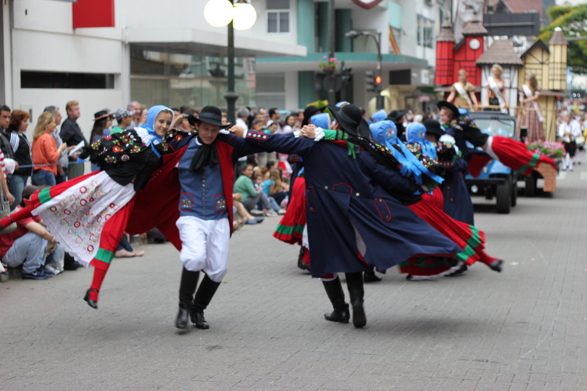 Grupo folclórico dançando de mãos dadas no desfile da Oktoberfest na Rua XV de Novembro, em Blumenau: mulheres de lenço azul, blusa bordada e saia vermelha giram com homens de chapéu preto e casaco azul, com público sentado na calçada e um carro alegórico de casinhas enxaimel ao fundo.