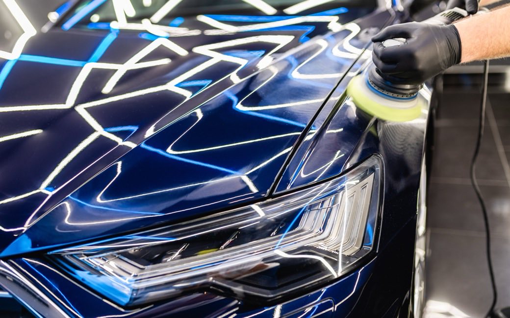 Profissional de oficina passando a politriz na lateral de um carro claro dentro de um box de estética automotiva iluminado.