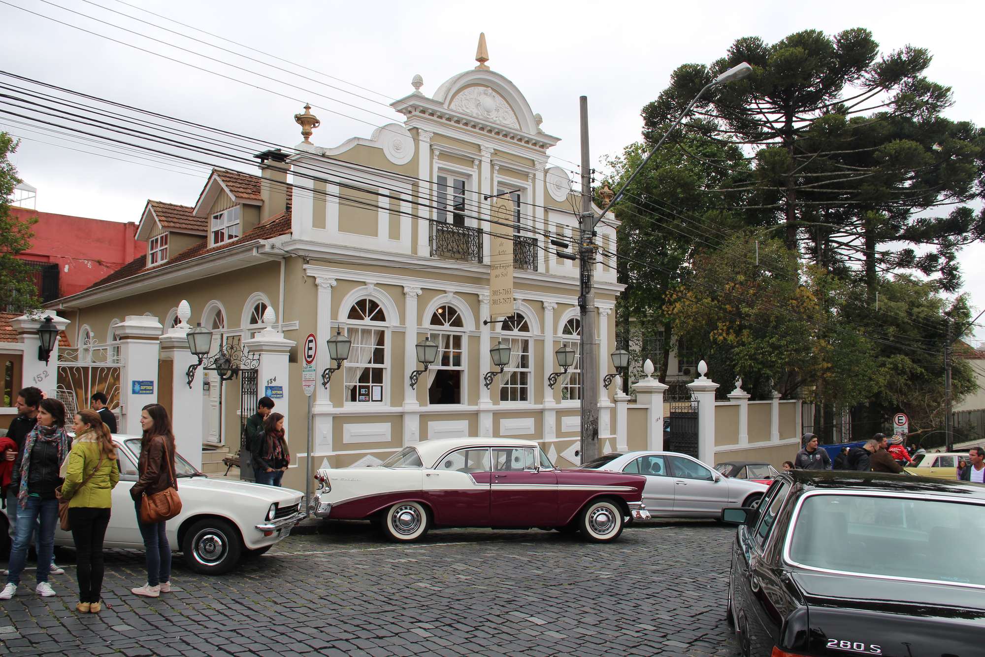 Exposição de carros antigos numa rua de paralelepípedos de Curitiba, com um Chevrolet Bel Air vinho e branco estacionado diante de um casarão histórico, visitantes conversando na calçada e araucárias ao fundo.
