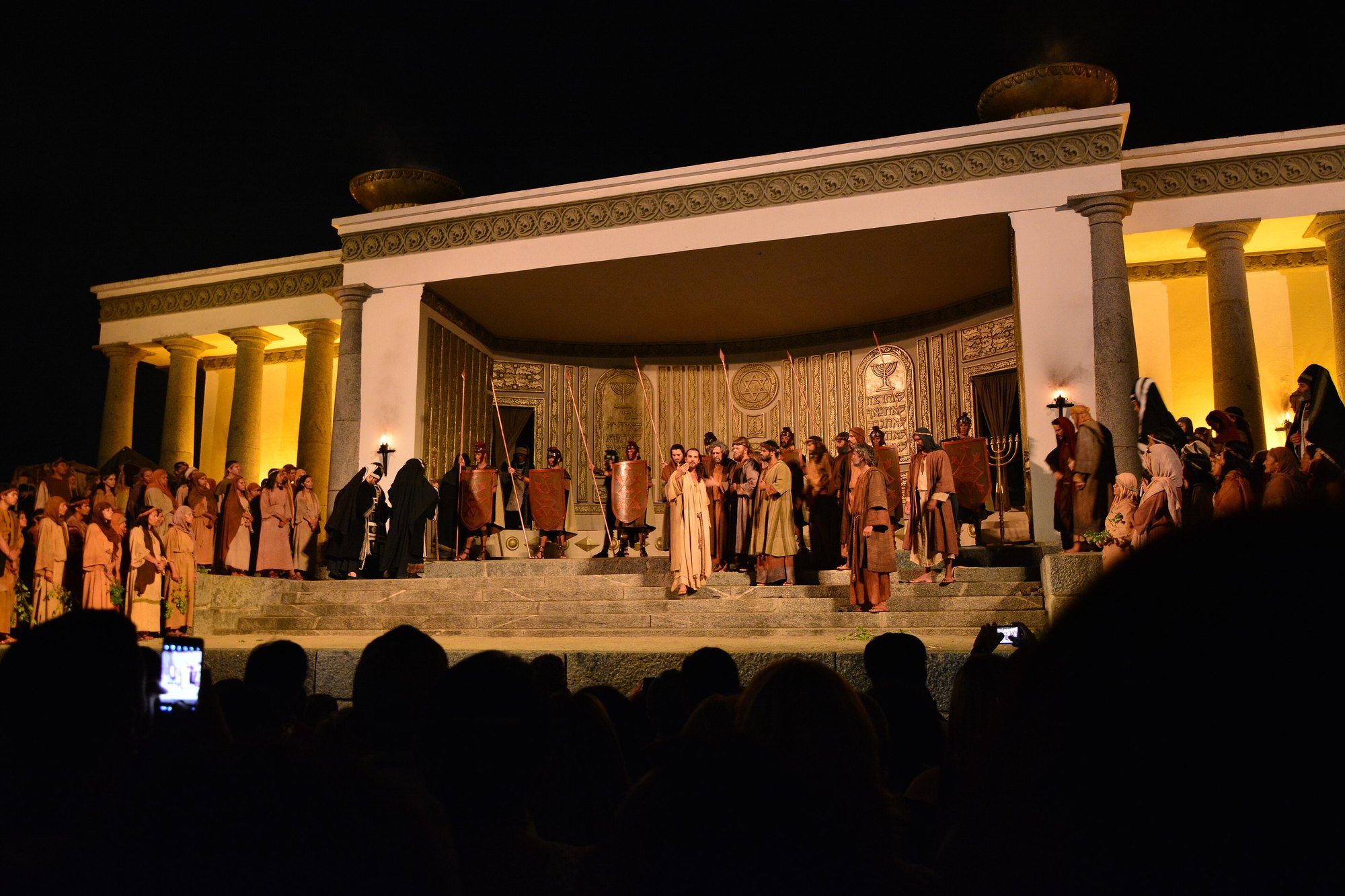 Cena da Paixão de Cristo no palco do Templo, em Nova Jerusalém, à noite: atores em trajes de época nas escadarias de um templo de colunas iluminado em amarelo, e a plateia em silhueta no primeiro plano, com dois celulares erguidos filmando.