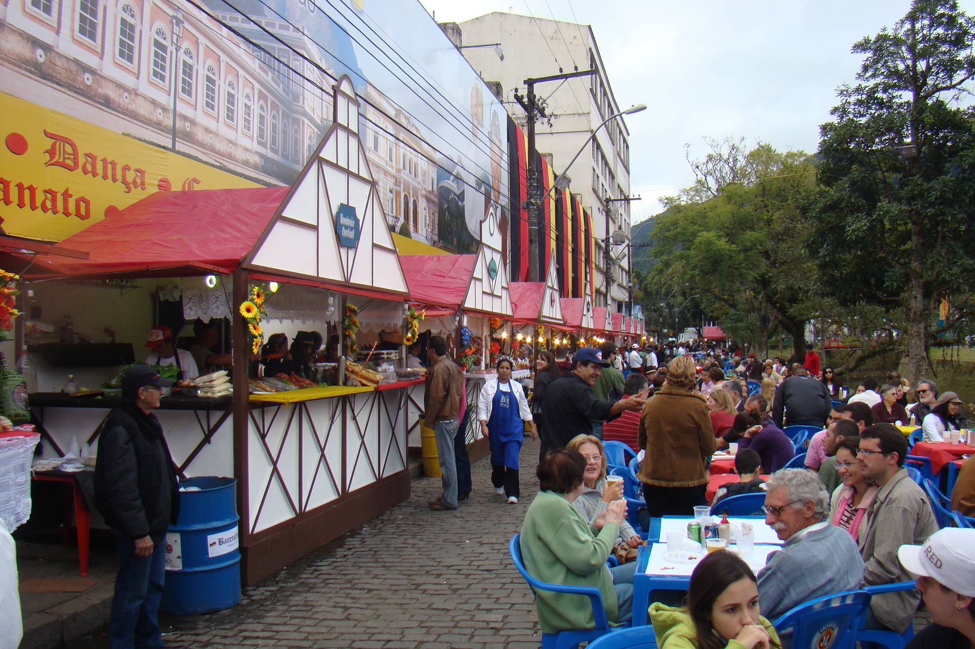Rua do centro de Petrópolis tomada por barracas de comida de madeira e mesas plásticas azuis cheias de gente comendo, com a mata da serra ao fundo, durante uma festa de rua da cidade.