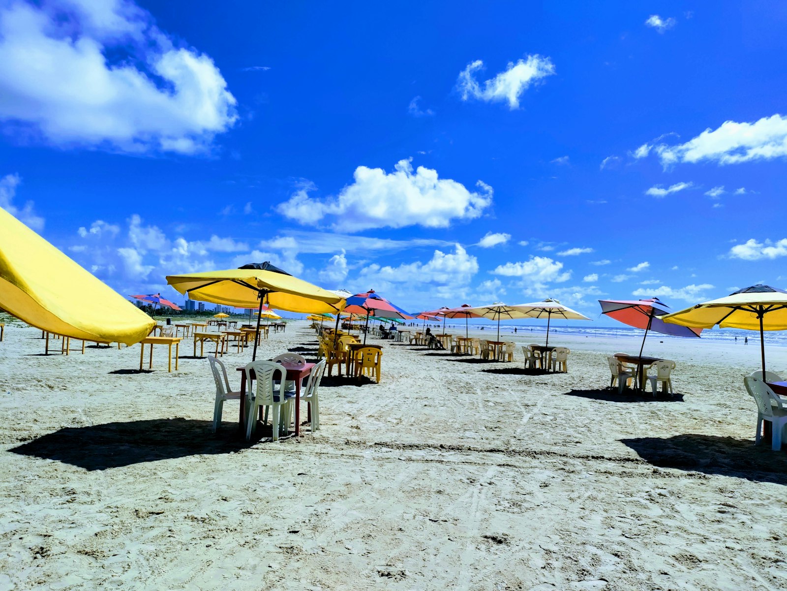 Fileira de guarda-sóis amarelos e mesas vazias na areia de uma praia em Aracaju, sob céu azul com nuvens.