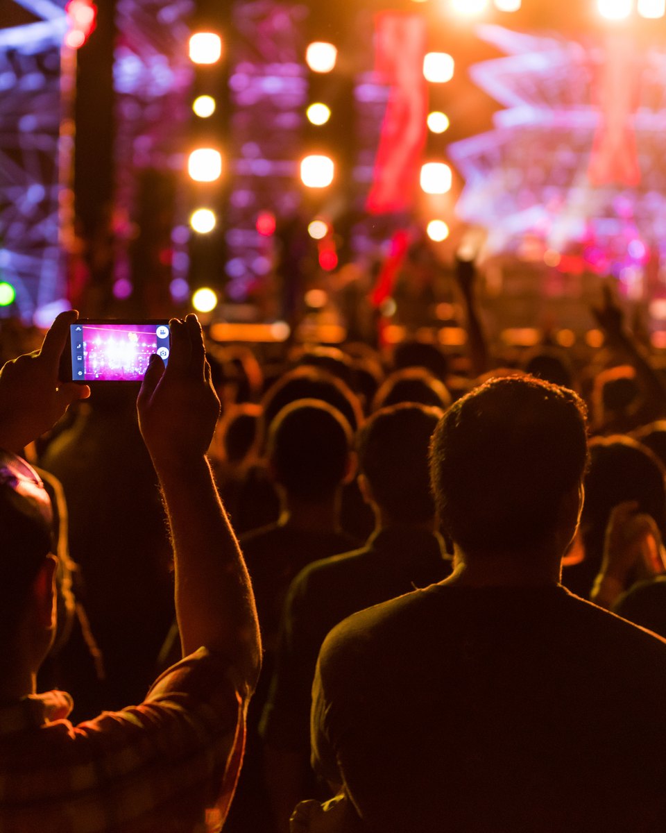 Plateia de costas num show à noite, com uma pessoa filmando o palco iluminado com o celular.
