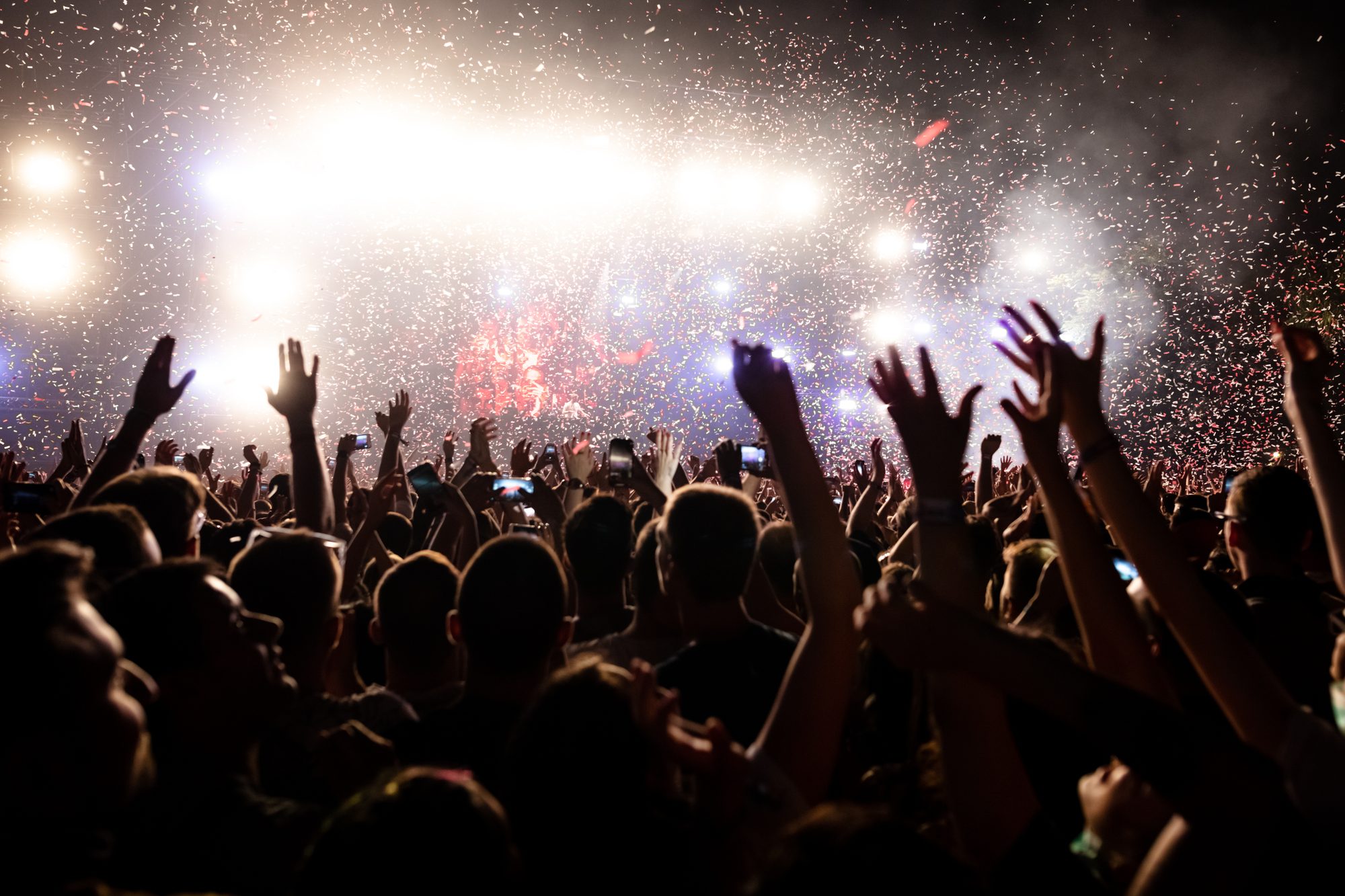 Multidão com os braços para cima sob chuva de confete e luzes do palco em um show à noite.
