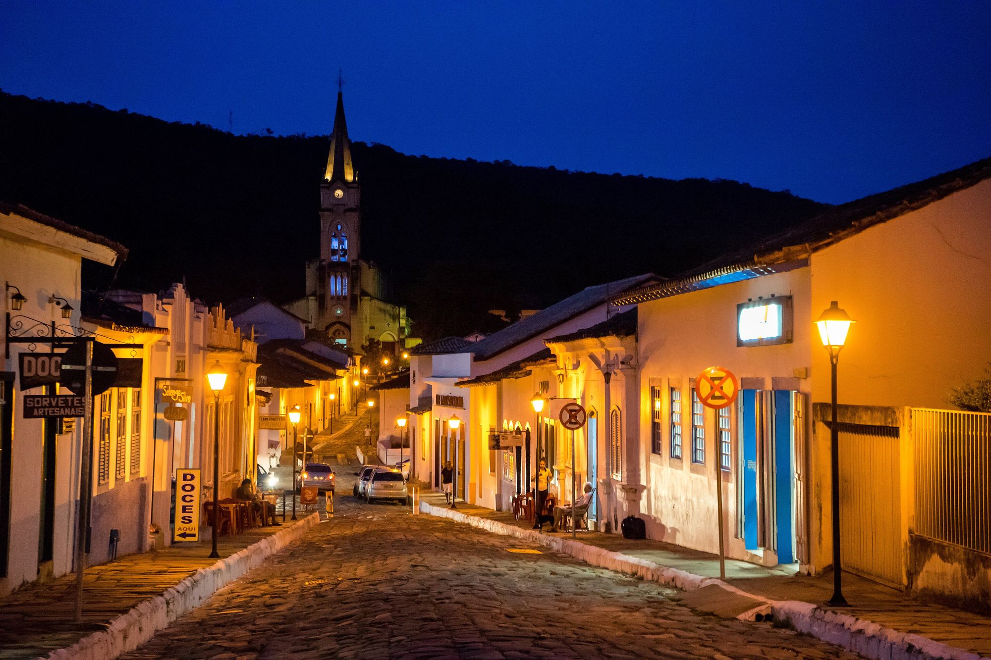 Rua de pedra do centro histórico da Cidade de Goiás ao anoitecer, com casarões coloniais iluminados por lampiões, placas de doces e sorvetes artesanais e a torre pontuda de uma igreja diante da serra, ao fundo.