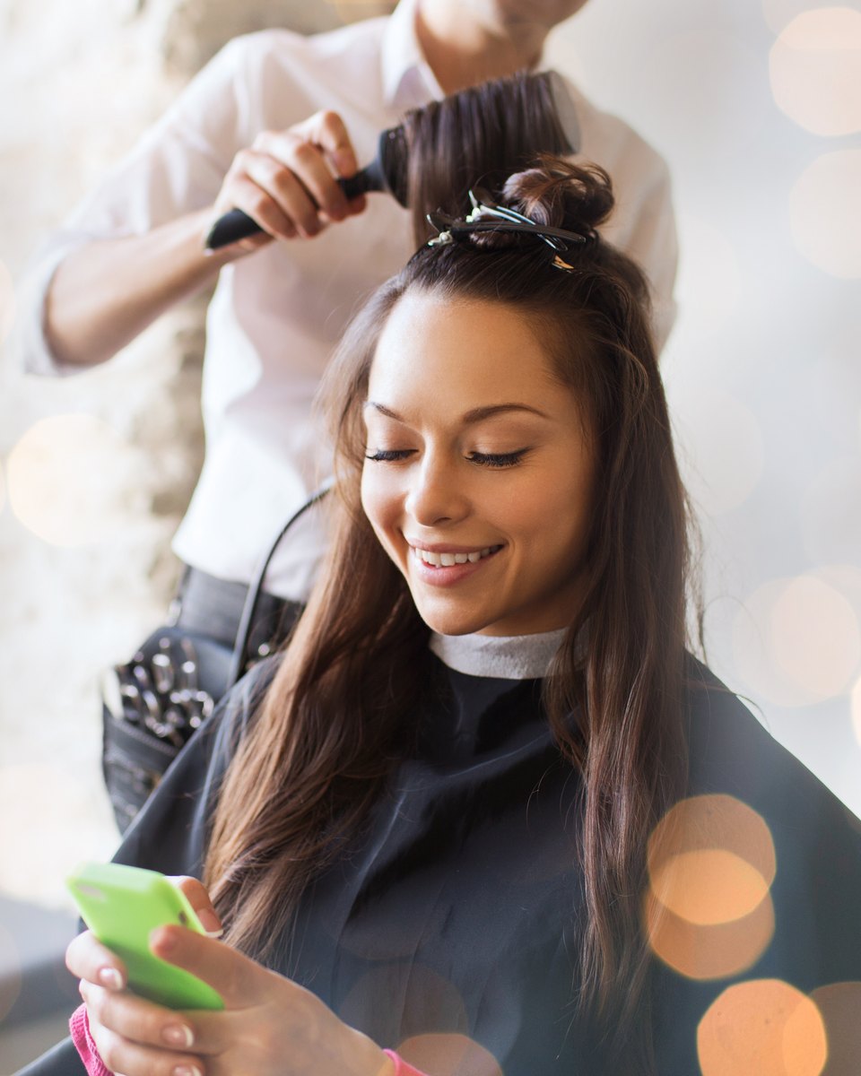 Jovem de cabelo longo castanho sorrindo para o celular verde na mão, sentada de capa preta enquanto a cabeleireira escova o cabelo dela num salão.