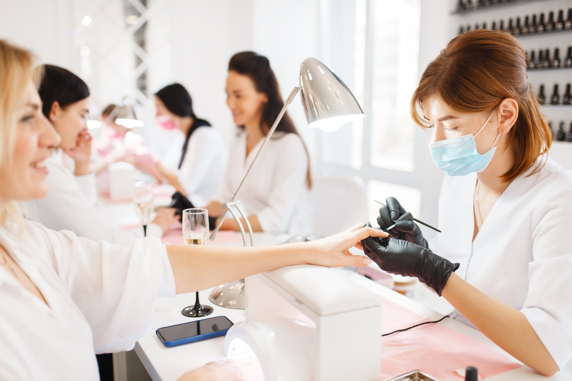 Manicure de máscara e luvas pretas pintando as unhas de uma cliente sorridente num salão claro, com outras clientes sendo atendidas nas mesas ao fundo.