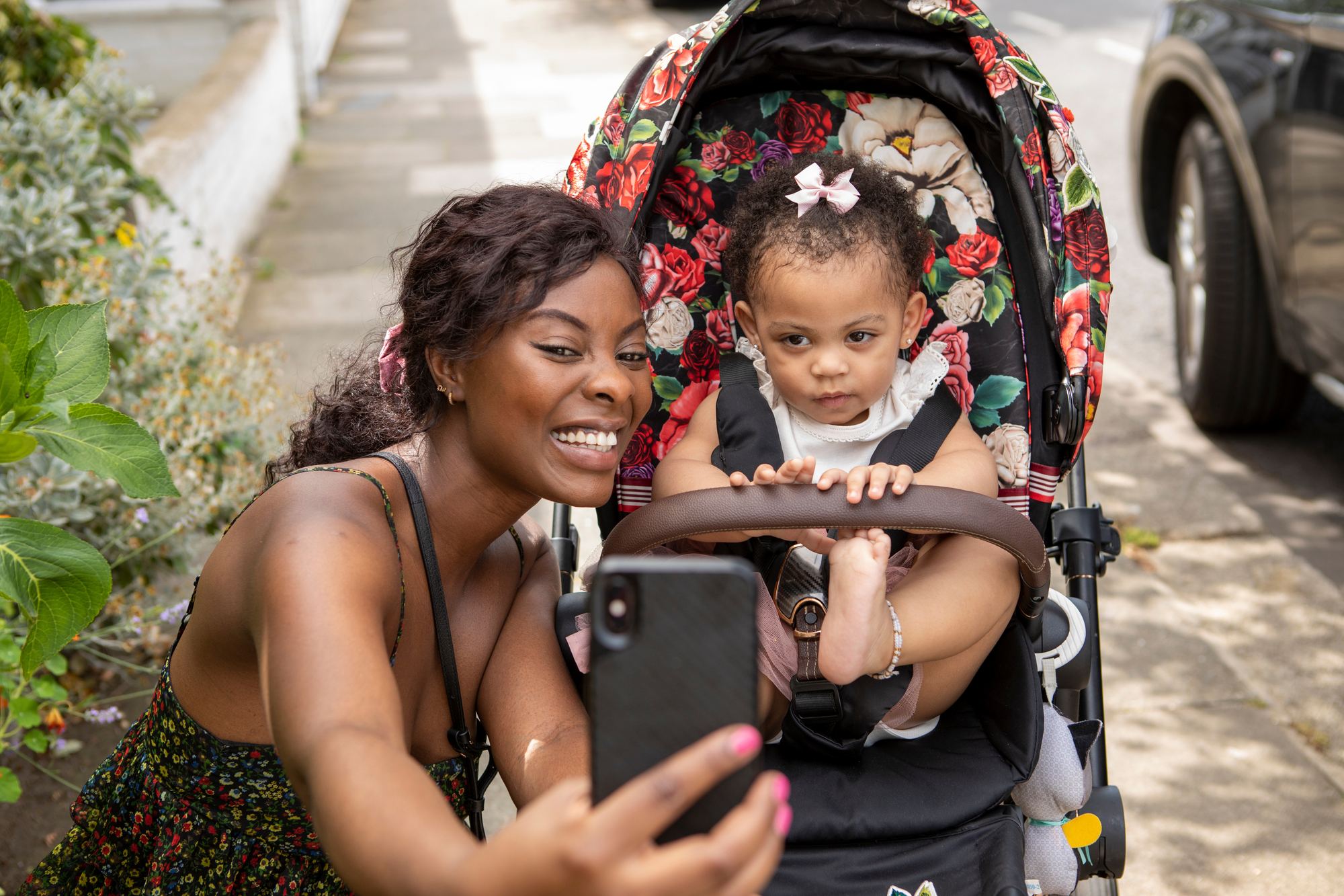 Mulher sorridente agachada na calçada tirando uma selfie com o celular ao lado da filha pequena, sentada num carrinho de bebê estampado de flores.