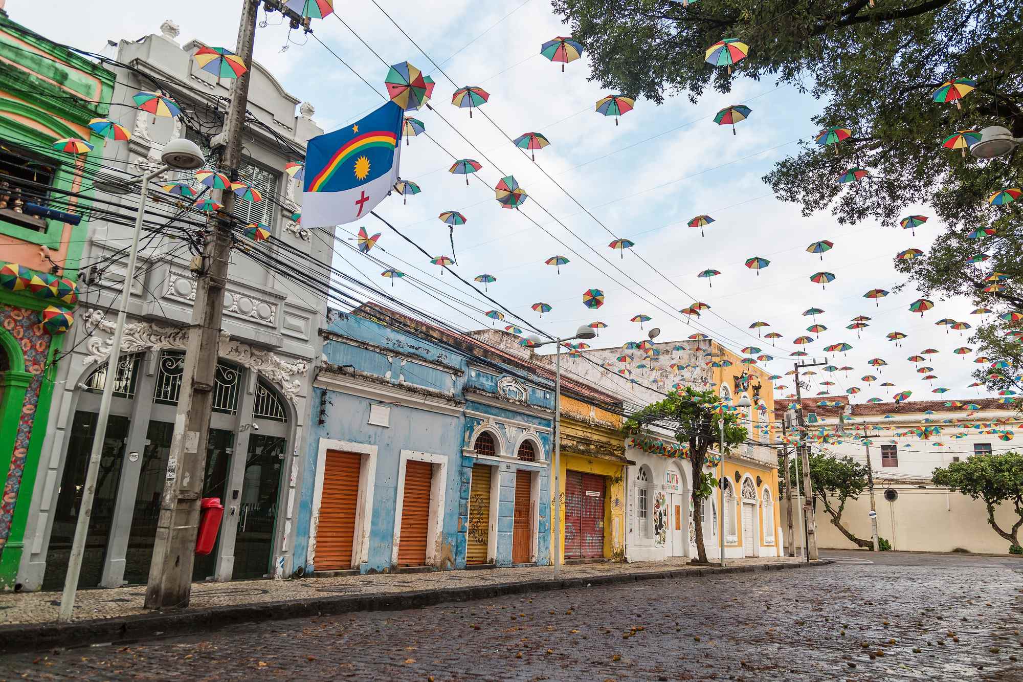 Rua de paralelepípedo do Bairro do Recife com casarões coloridos, sombrinhas de frevo penduradas nos fios e a bandeira de Pernambuco num poste, no Recife Antigo.