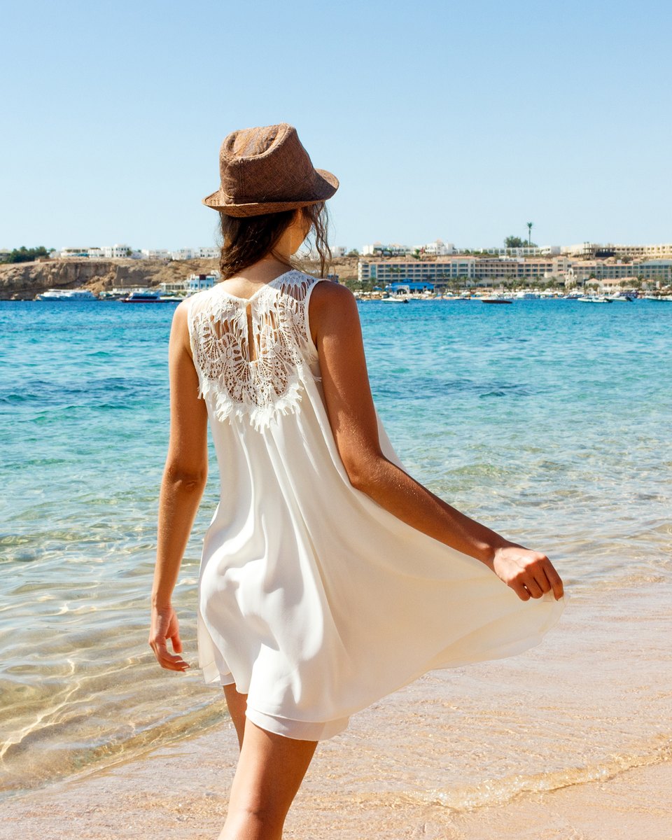 Mulher de vestido branco curto com renda nas costas e chapéu de palha, de costas, andando na beira do mar numa praia de água clara, num dia de sol.