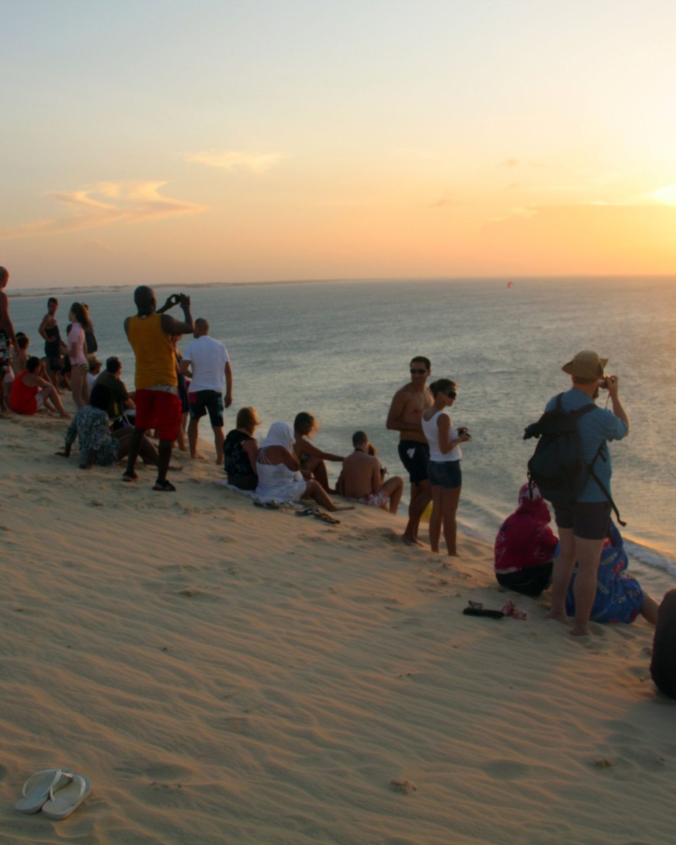 Pessoas sentadas e em pé no alto da Duna do Pôr do Sol, em Jericoacoara, fotografando o sol que se põe sobre o mar, com a areia dourada em primeiro plano.