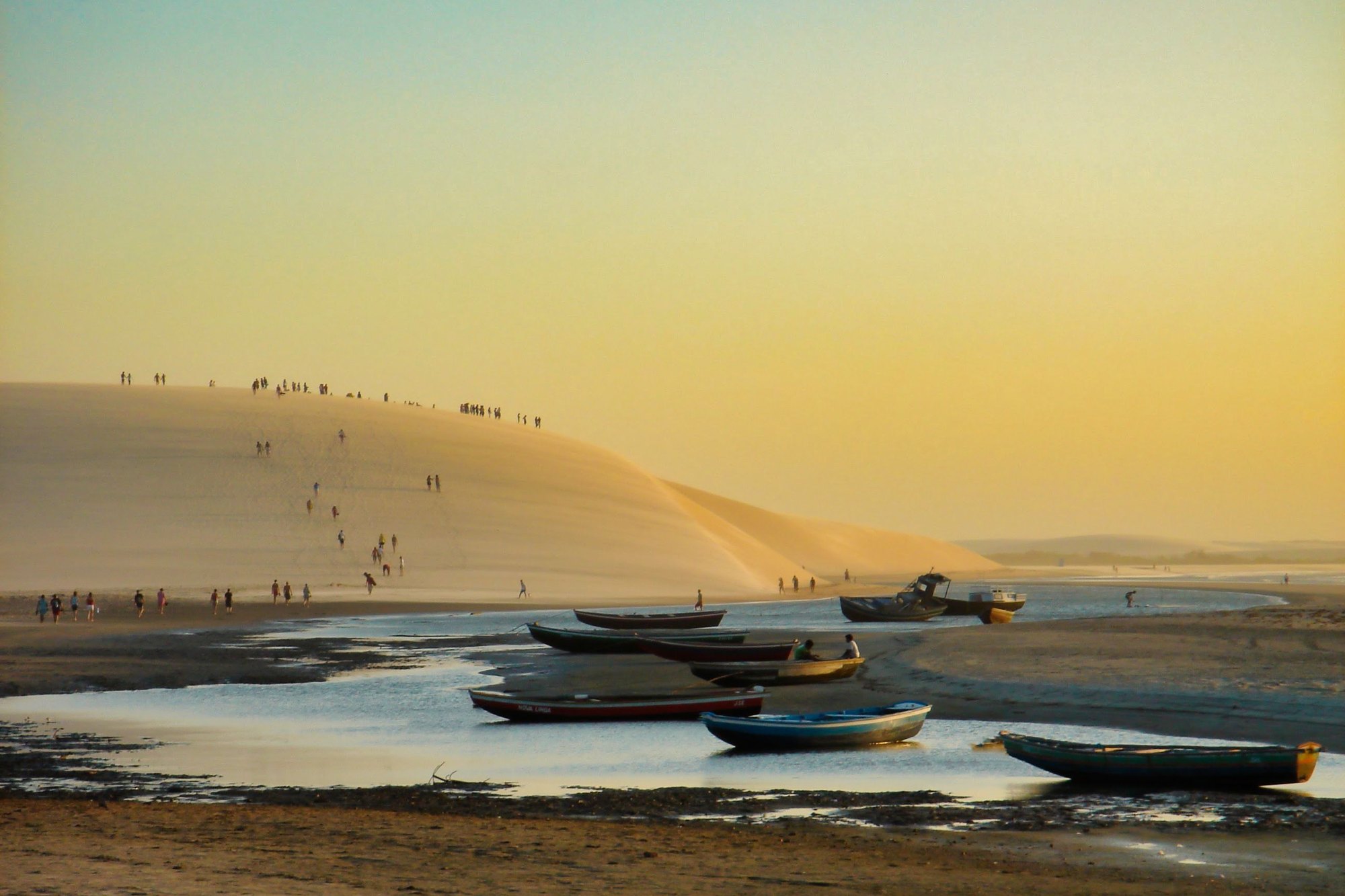Fim de tarde dourado em Jericoacoara, com dezenas de pessoas subindo e caminhando no alto da Duna do Pôr do Sol e barcos de pesca parados na água rasa da maré baixa, em primeiro plano.