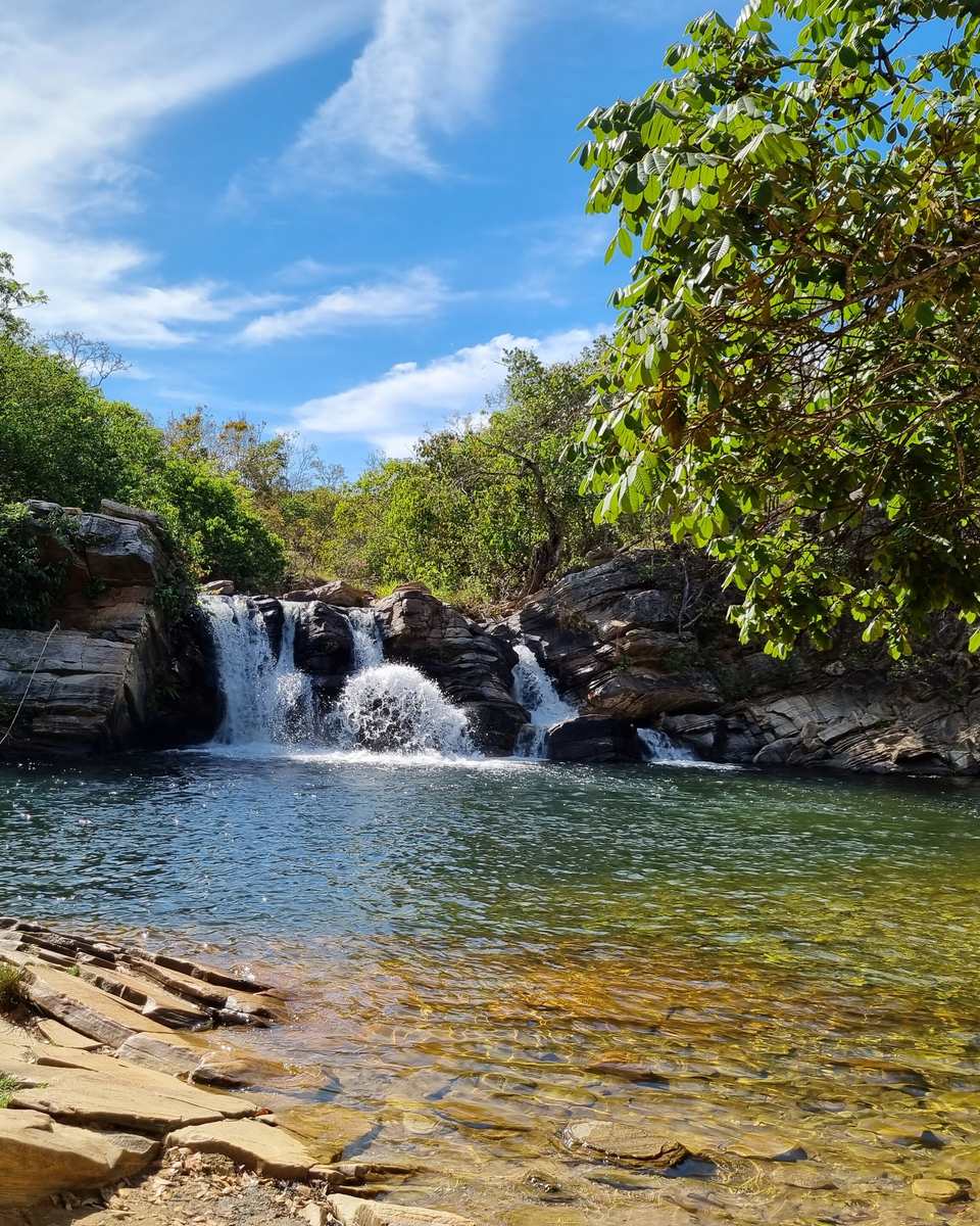 Duas pessoas nadando no poço de água cristalina da Cachoeira das Araras, em Pirenópolis, num dia de céu azul, com a queda d'água entre pedras e a mata do Cerrado em volta.