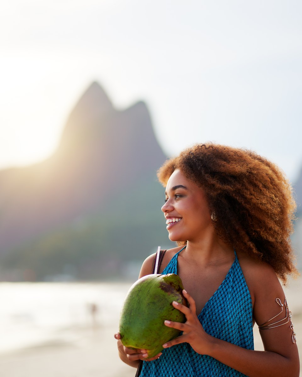 Mulher de cabelo cacheado e regata azul sorrindo com um coco verde nas mãos na areia de Ipanema, no fim da tarde, com o Morro Dois Irmãos ao fundo.
