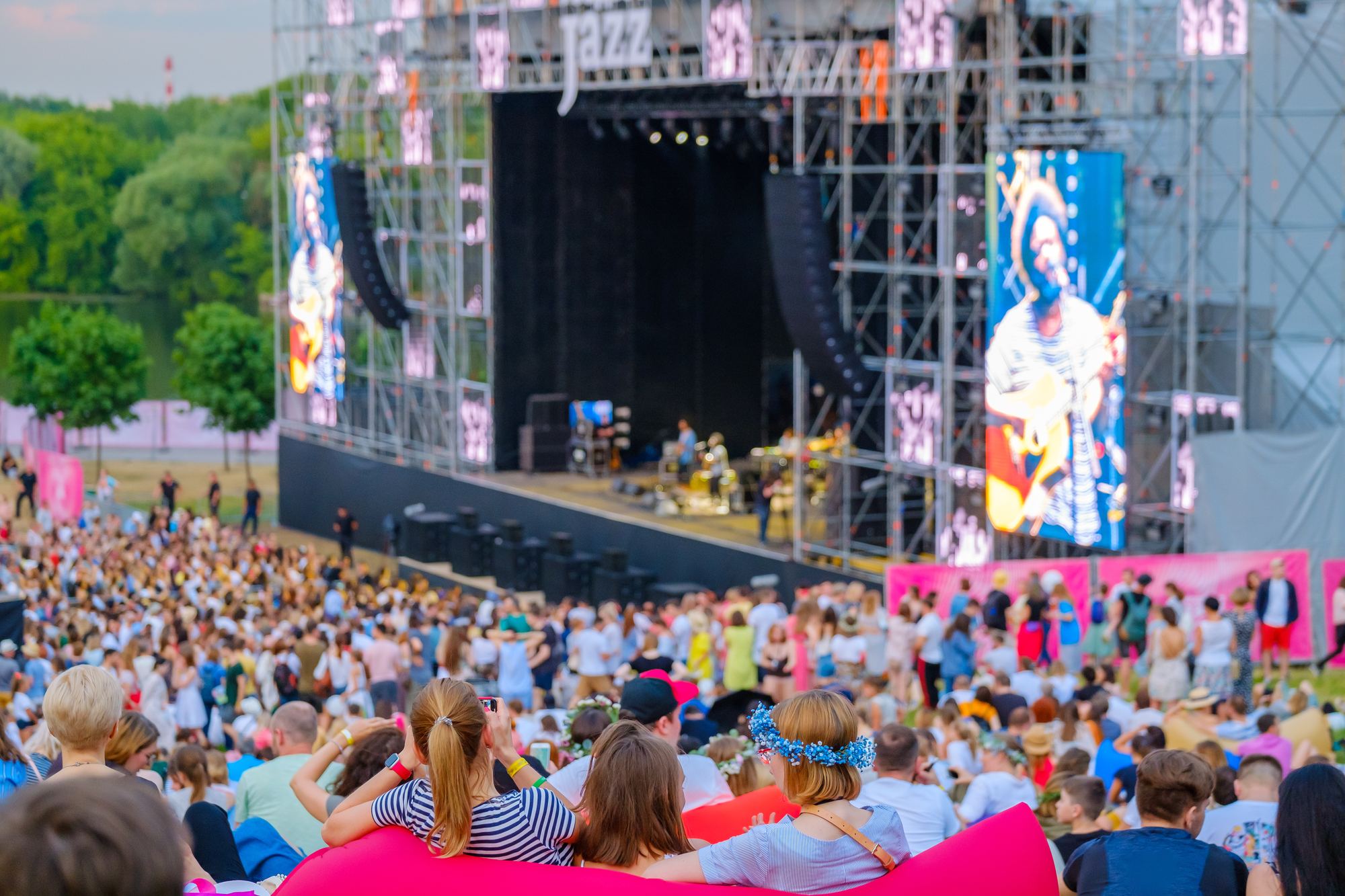 Público sentado e em pé na grama diante de um palco ao ar livre, de dia, com árvores ao fundo e duas amigas num pufe rosa em primeiro plano.