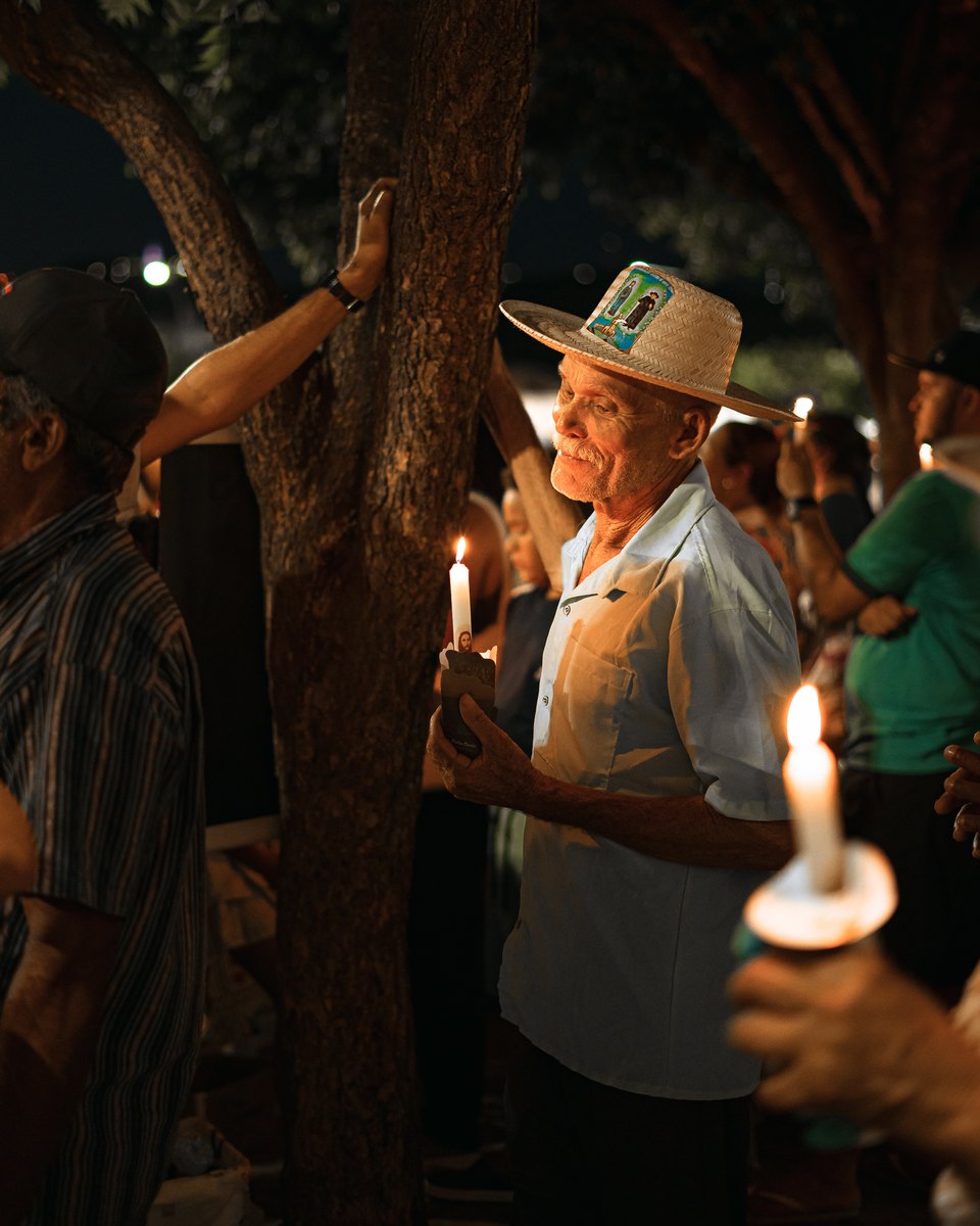 Romeiro idoso de chapéu de palha enfeitado, segurando uma vela acesa entre outros fiéis, à noite, durante o cortejo de Nossa Senhora das Candeias, em Juazeiro do Norte.
