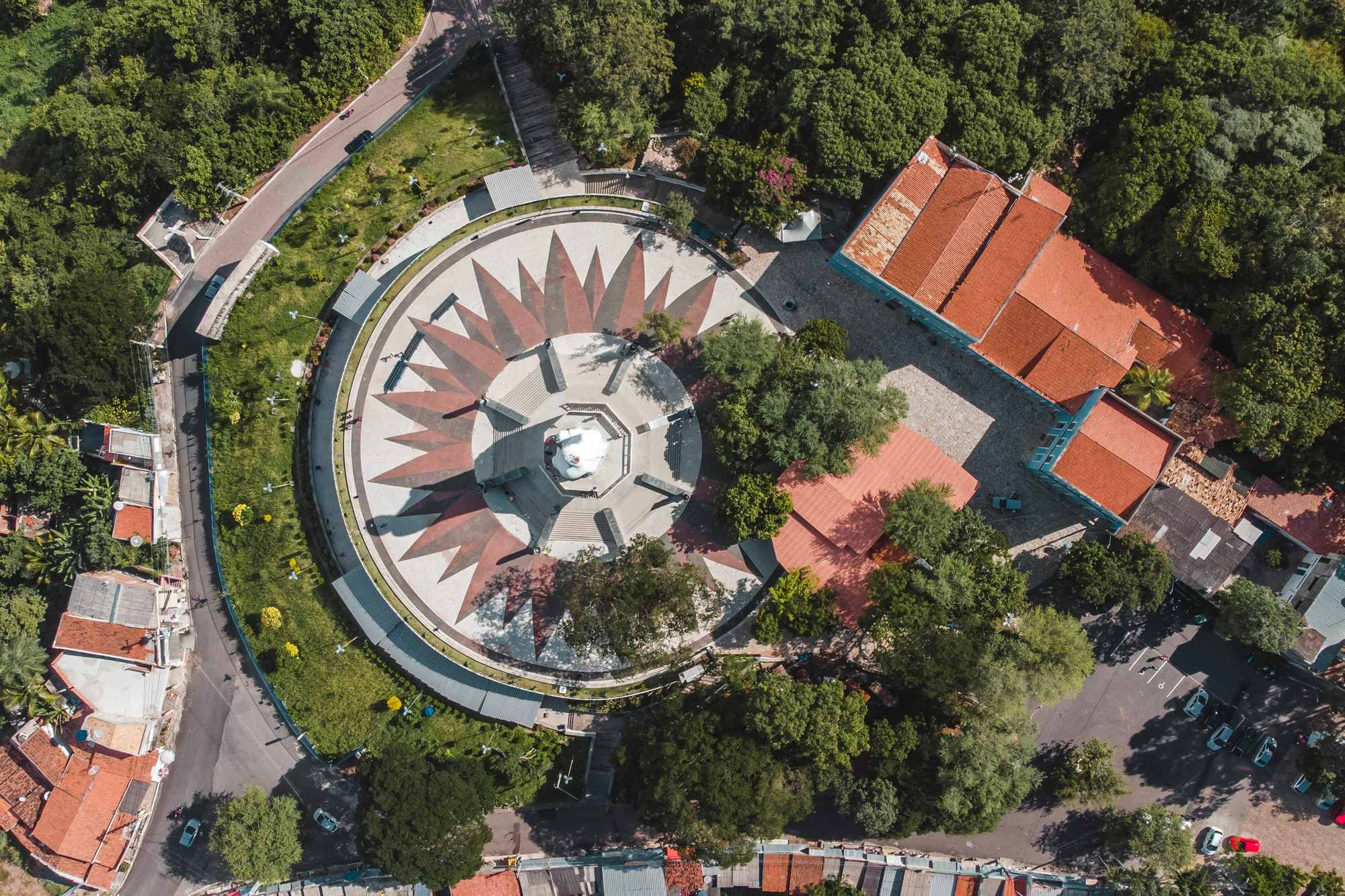 Vista aérea, de cima, da praça circular da Colina do Horto, em Juazeiro do Norte, com a estátua branca do Padre Cícero no centro, o piso em estrela, casas de telhado vermelho e a mata da serra em volta.