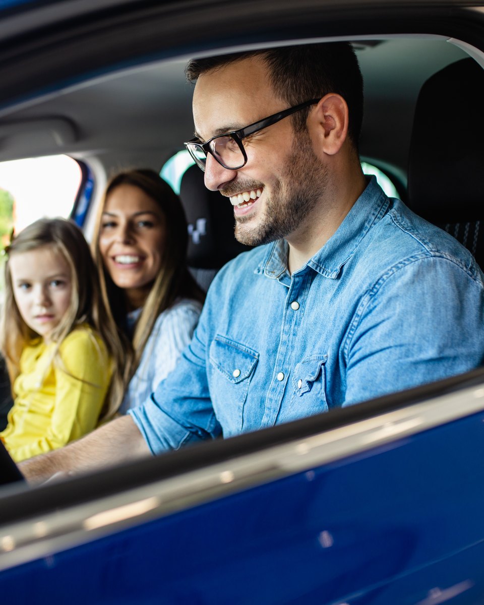 Homem de óculos e camisa jeans sorrindo ao volante de um carro azul numa loja de carros, com uma mulher e uma menina no banco de trás.