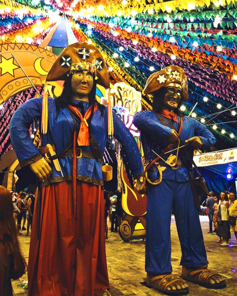 Esculturas gigantes de Maria Bonita e Lampião, com chapéus de couro estrelados e roupas azuis, sob um teto de bandeirinhas coloridas e cordões de luz no arraial do São João de Cruz das Almas, no Recôncavo baiano, com o público passeando ao fundo.