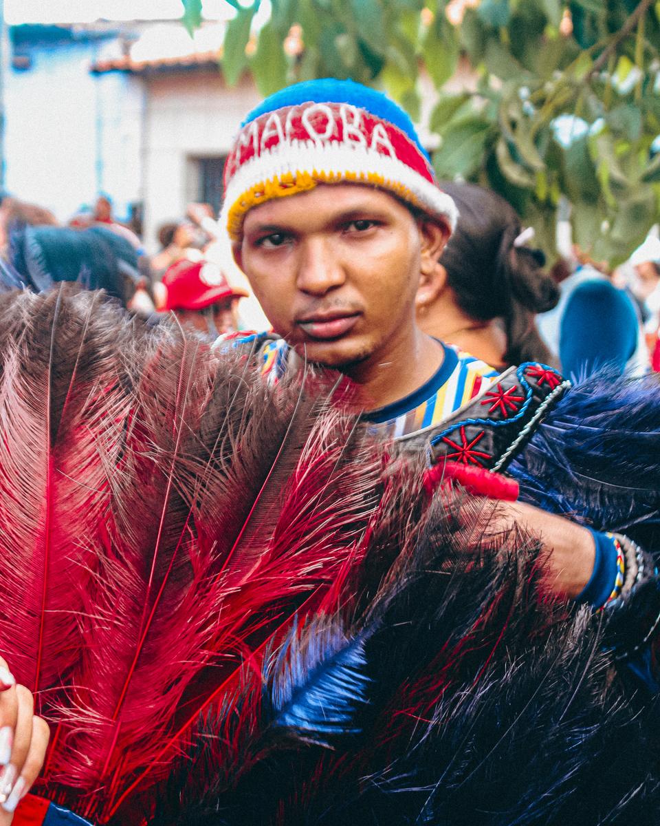 Caboclo de pena do Boi da Maioba segurando um leque de plumas vermelhas e azuis, de gorro bordado e peitoral de veludo, em meio à multidão numa rua de São Luís.
