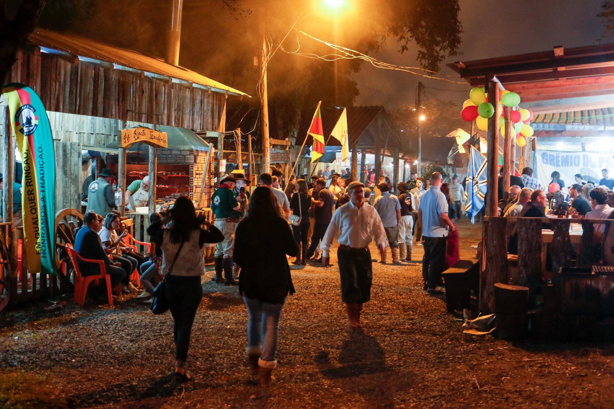 Rua de chão batido entre piquetes de madeira no Acampamento Farroupilha, no Parque Harmonia, em Porto Alegre, à noite, com gaúchos de bombacha e visitantes caminhando, bandeiras do Rio Grande do Sul e gente sentada em volta da churrasqueira.