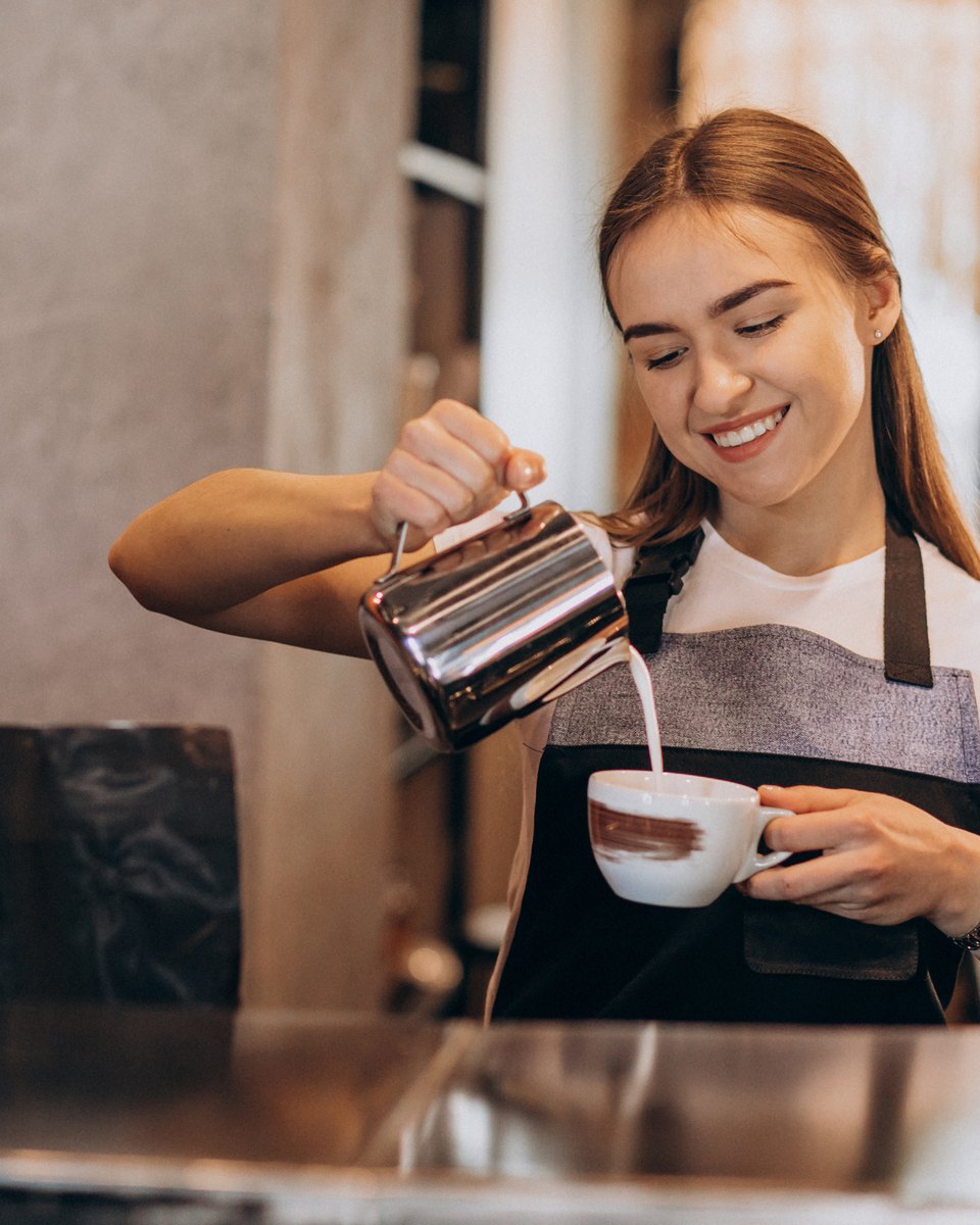 Barista sorridente de avental preto despejando leite vaporizado numa xícara no balcão de uma cafeteria, com a máquina de espresso ao lado.