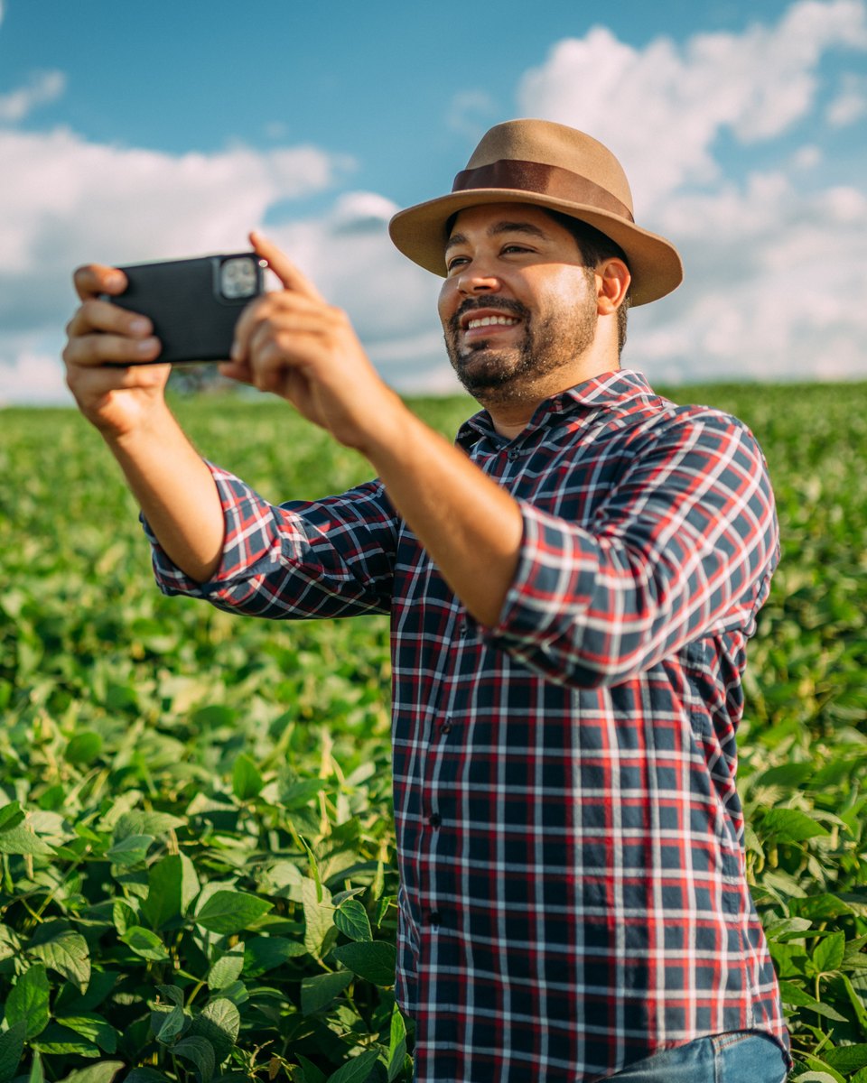 Produtor de chapéu e camisa xadrez sorrindo enquanto filma a lavoura de soja com o celular, em pé no meio do talhão.
